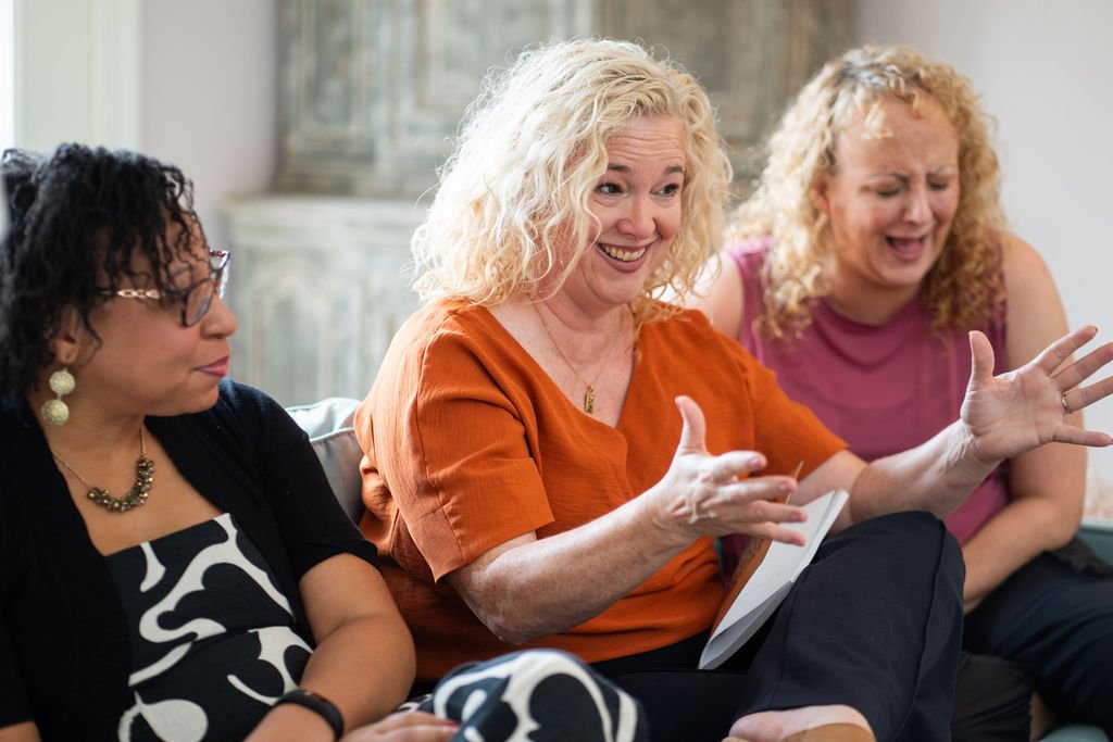 Three women sitting on a couch and engaging in a conversation; the woman in the middle appears to be speaking animatedly.