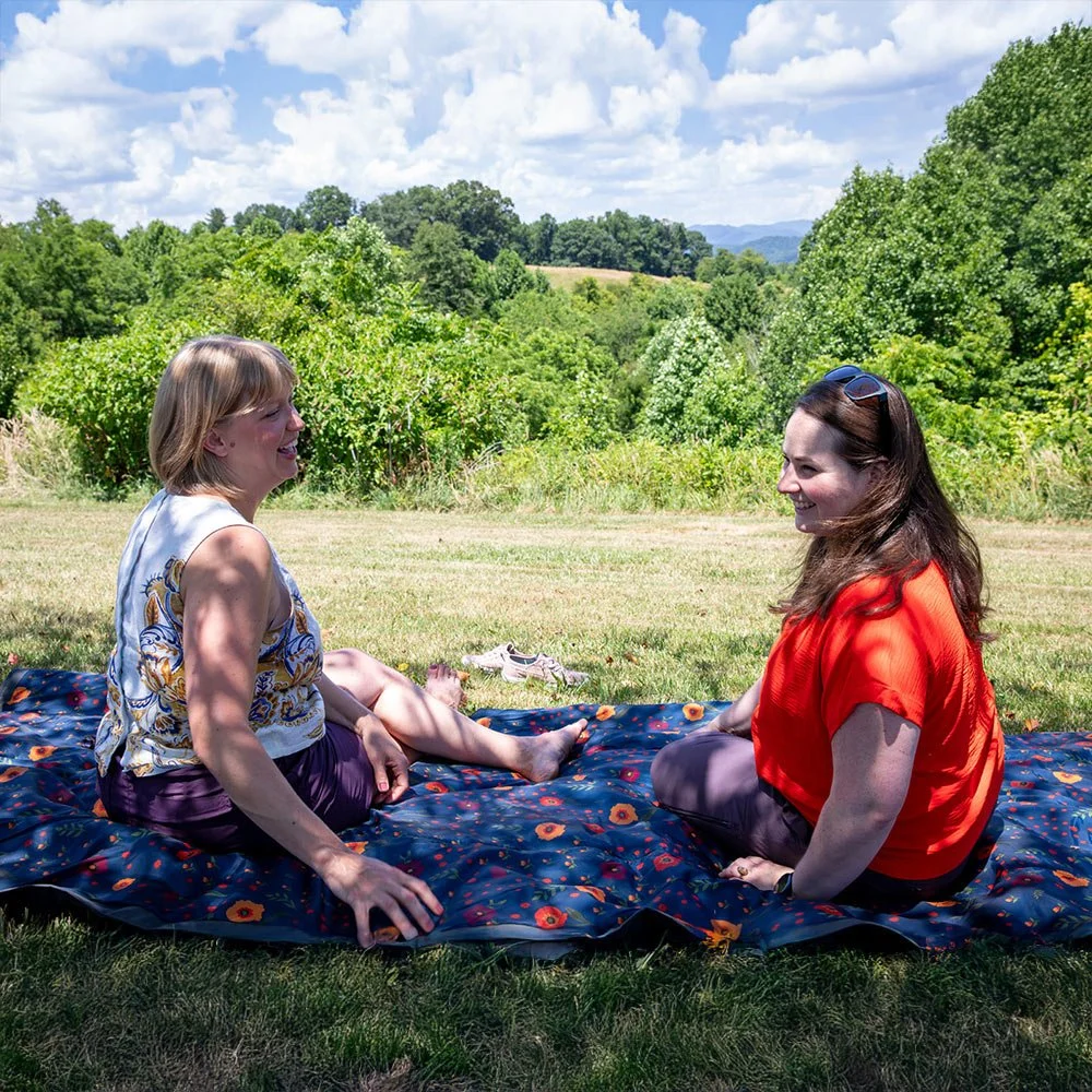 Two women sitting on a blanket on grass, laughing and facing each other, with a background of trees and a partly cloudy sky.
