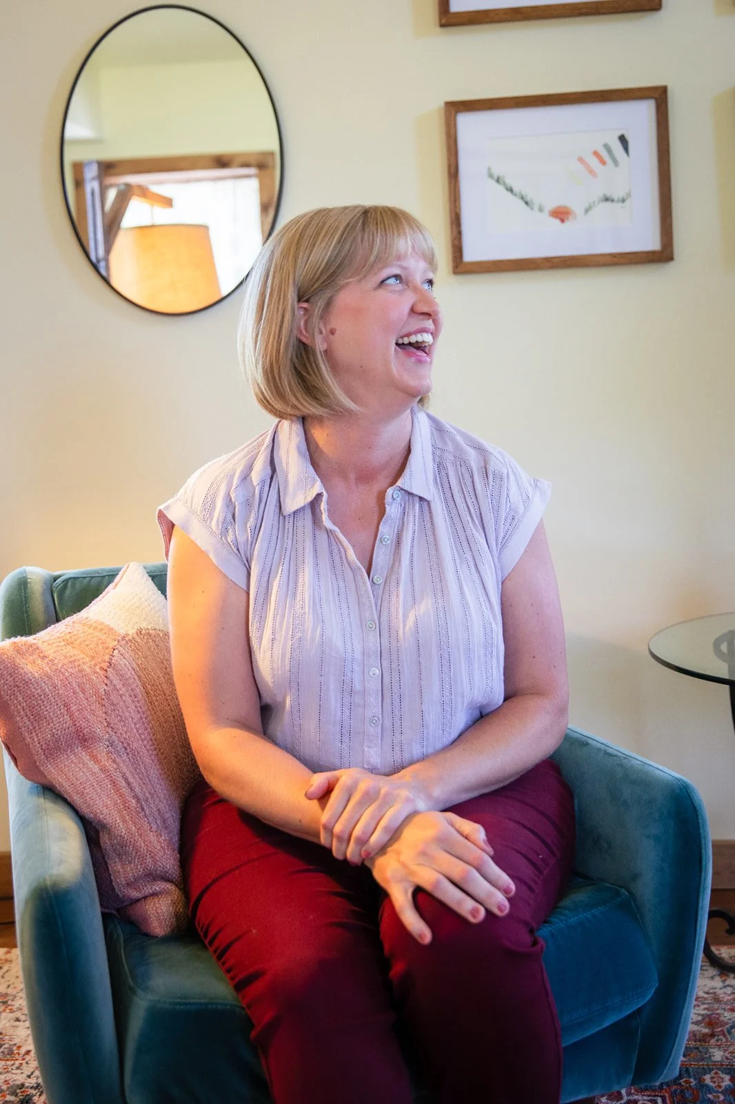 A woman with blonde hair laughing and smiling while sitting on a teal armchair in a cozy room. Behind her, a round mirror and framed artwork are on the wall.