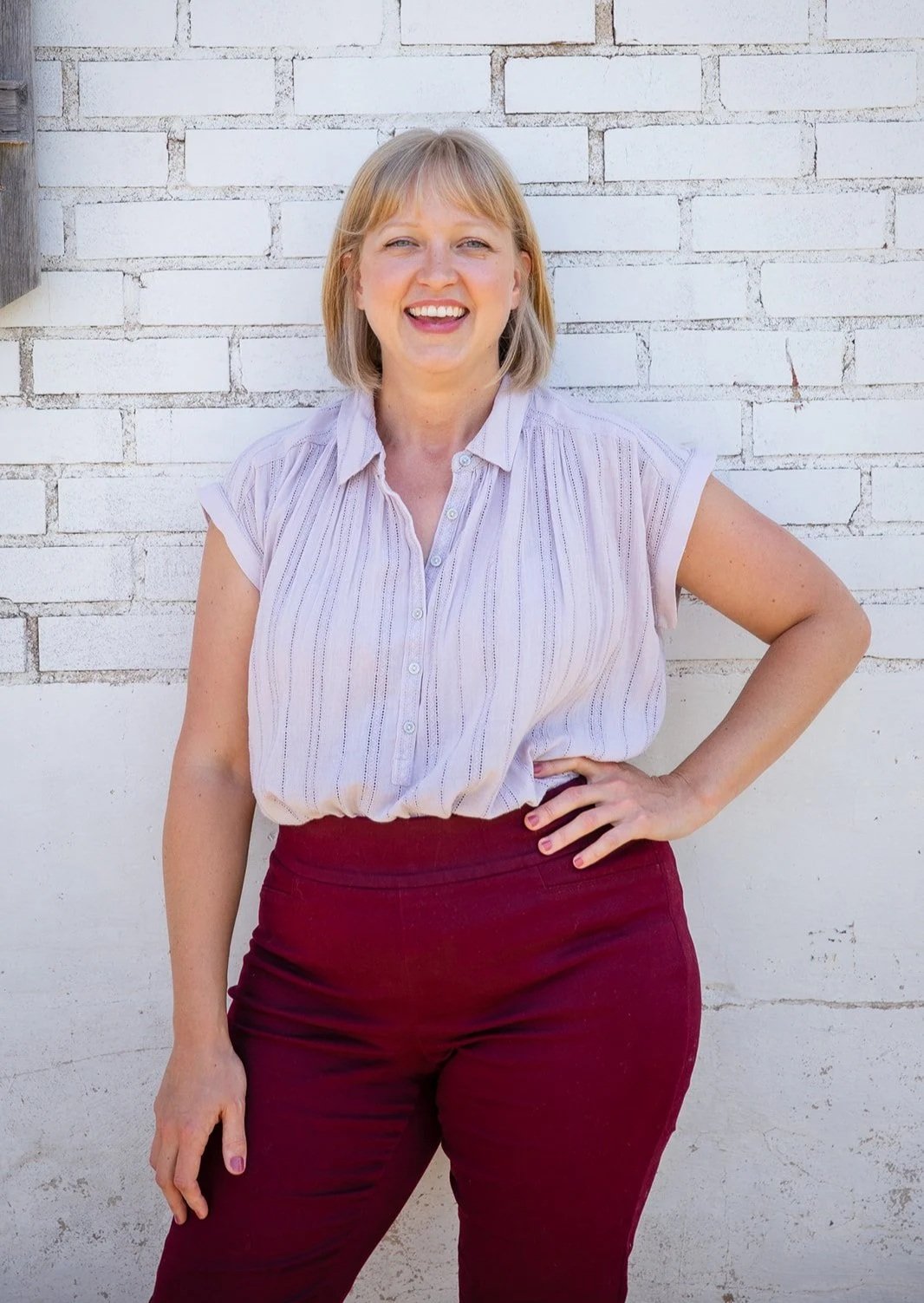 A woman with blonde hair smiling, standing outdoors against a white brick wall, wearing a light-colored short-sleeve button-up shirt and maroon pants.