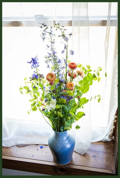 A colorful bouquet of flowers in a blue vase on a wooden windowsill with sheer curtains and natural light.