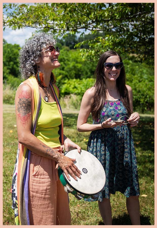 Two women outdoors, one with gray curly hair and tattoos, playing a tambourine, and the other with long dark hair, sunglasses, and a colorful dress, smiling under a tree.
