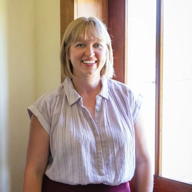 A smiling woman with blonde hair standing in front of a wooden window frame, wearing a short-sleeved, light-colored, striped blouse.