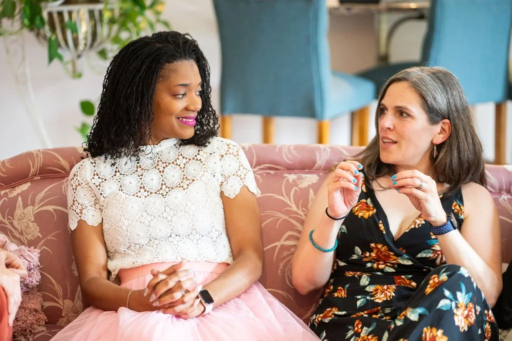 Two women sitting on a pink floral-patterned couch having a conversation indoors.