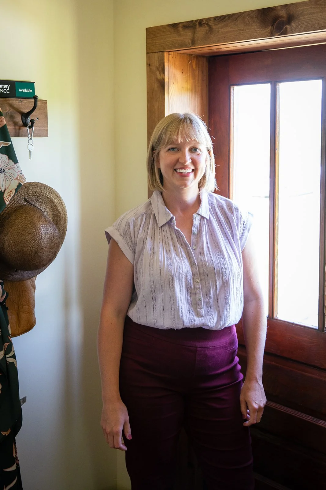 A smiling woman standing indoors near a wooden window, wearing a light purple striped shirt and maroon pants.