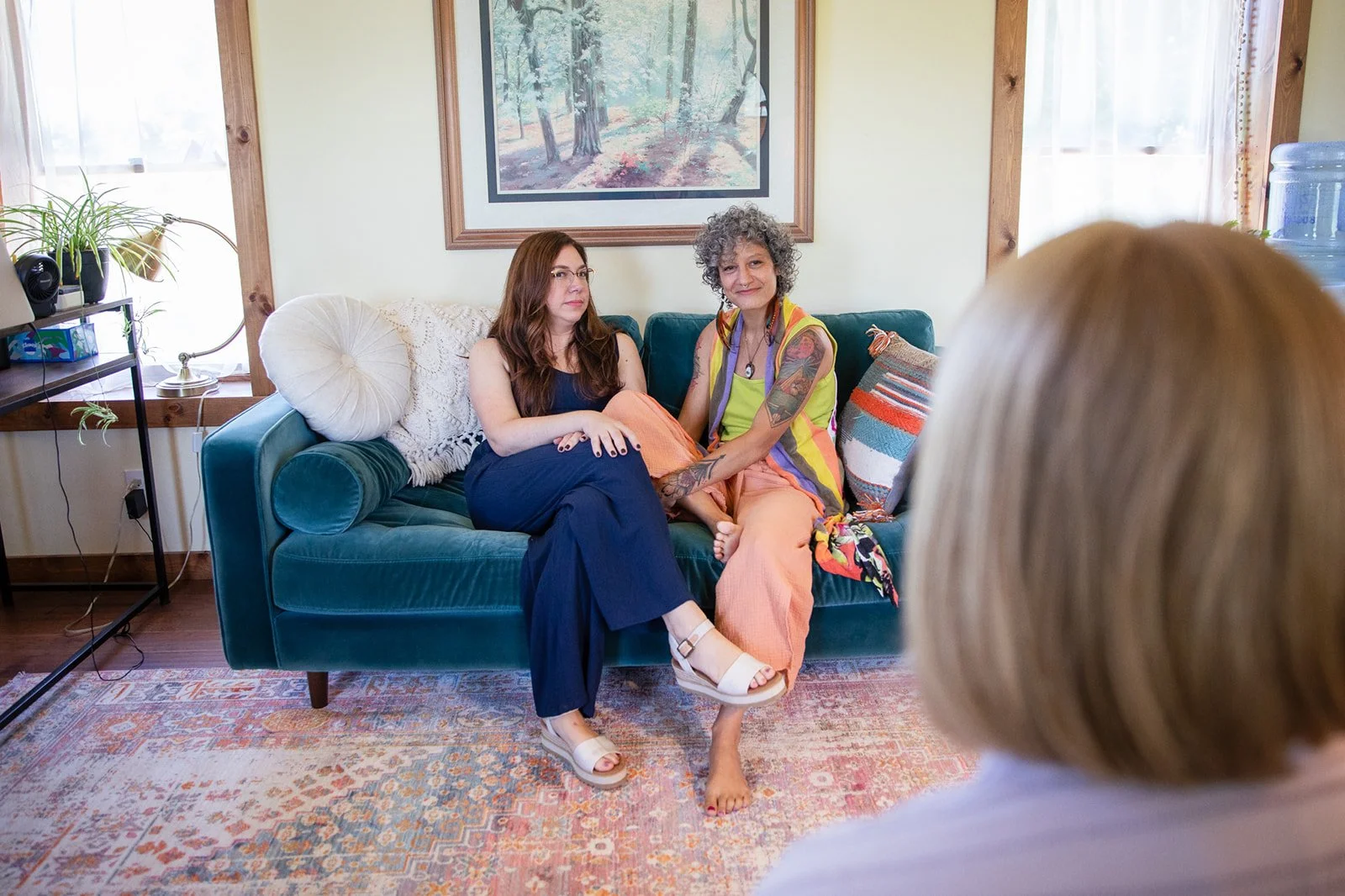 Two women sitting on a teal velvet couch in a living room, holding hands, with a third person's head visible in the foreground. One woman has long brown hair, glasses, a sleeveless navy dress, and white sandals. The other woman has short gray curly hair, tattoos, a sleeveless multicolored top, and peach pants. A painting of trees hangs on the wall behind them, and there are windows with curtains and potted plants nearby.
