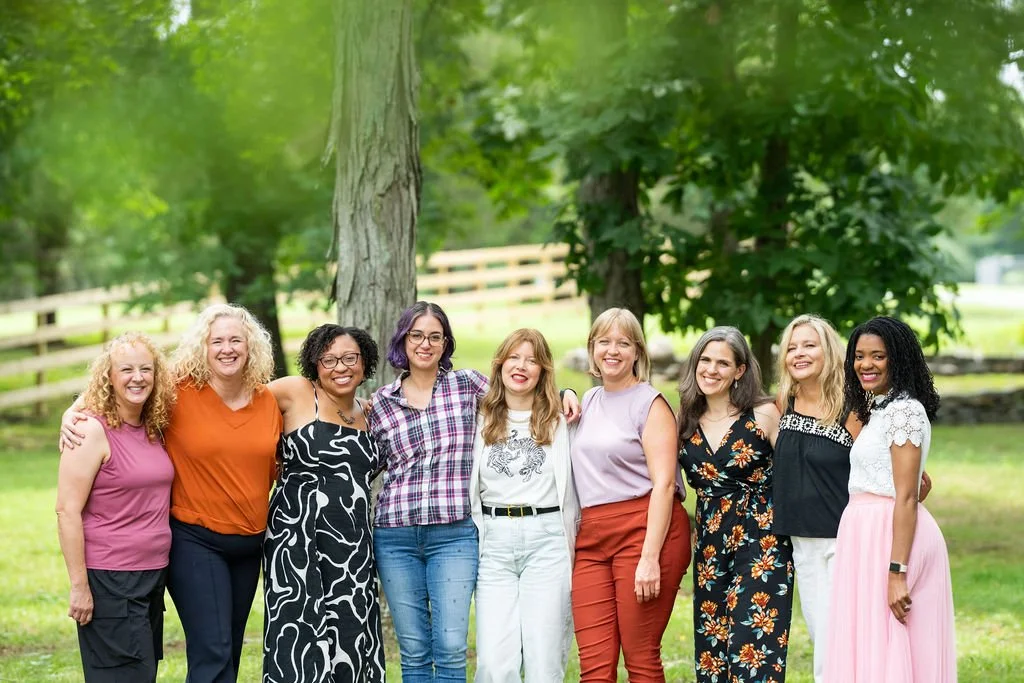 A group of nine women standing outdoors under a large tree, smiling, with a wooden fence in the background.