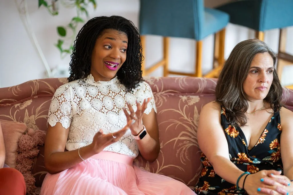 Two women sitting on a pink floral-patterned couch, one is smiling and talking, the other looks serious, in a well-lit room with green plants in the background.