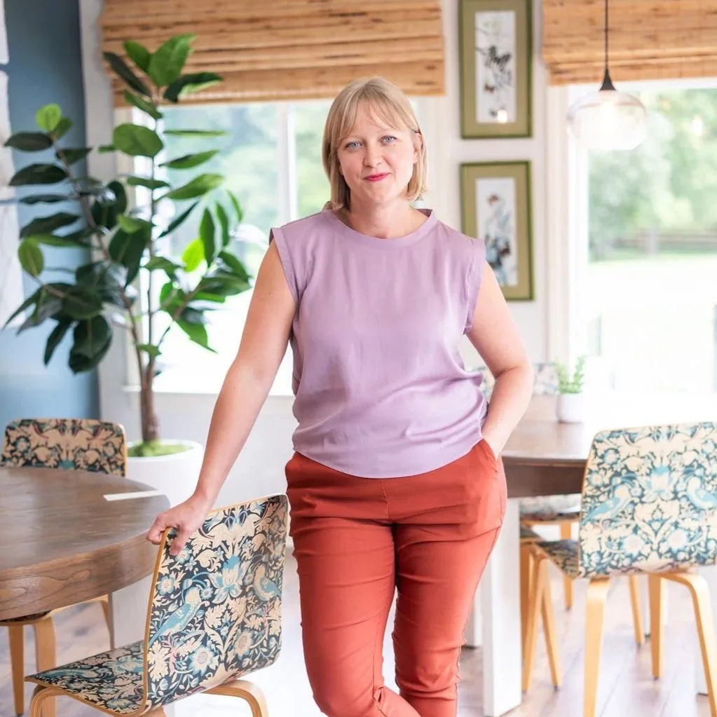 A woman standing in a bright room with wooden walls, plants, and framed pictures, wearing a purple sleeveless top and orange pants.
