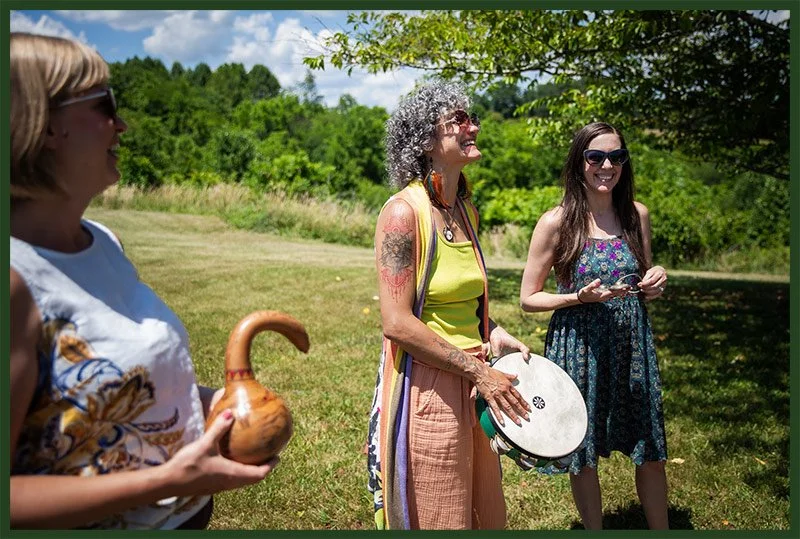 Three women outdoors on a sunny day, one holding a gourd-shaped instrument, another playing percussion with a tambourine, and the third smiling and holding a small object, surrounded by green trees and grass.