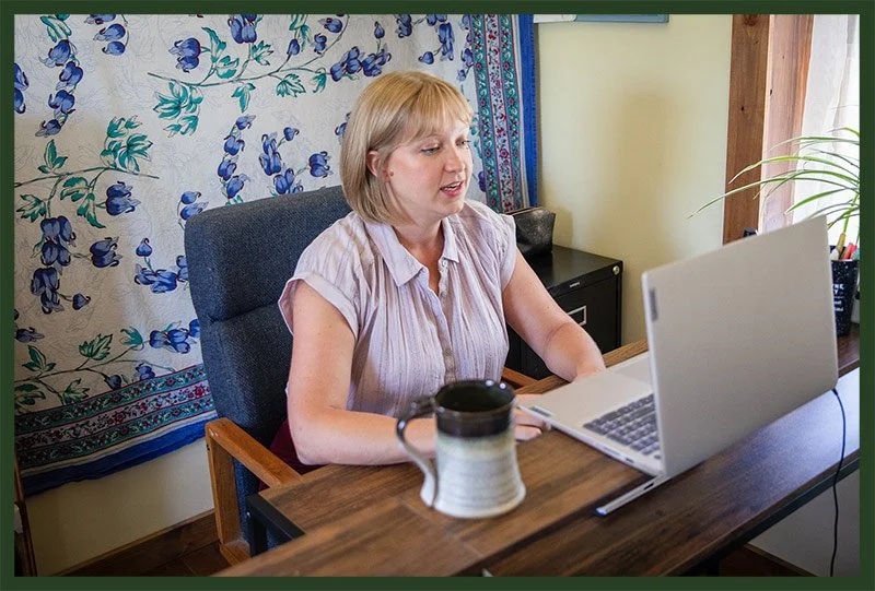 A woman with blonde hair providing online therapy on a silver laptop at a wooden desk, with a coffee mug nearby, in a room decorated with a floral tapestry and a potted plant.