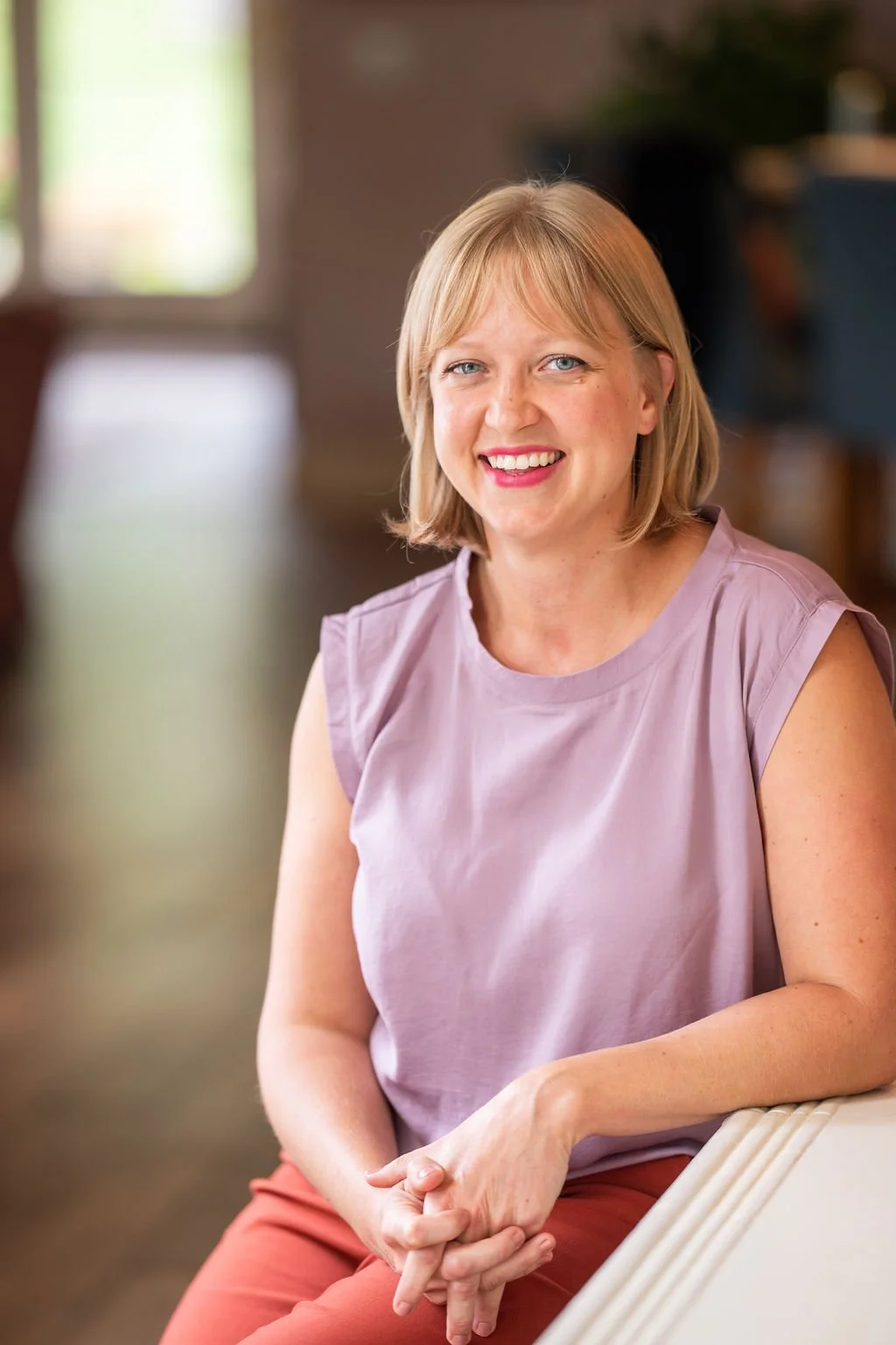 A woman with blonde hair and a pink sleeveless top smiling while sitting indoors with a blurred background.