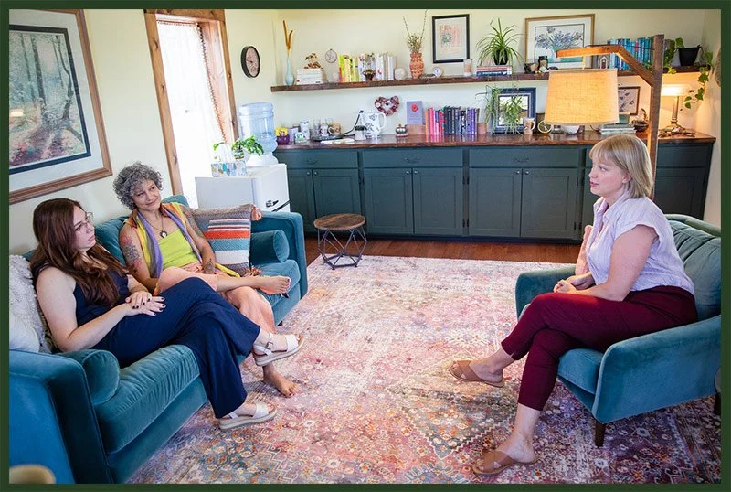 A couples therapy session in a cozy living room near Asheville NC with two women sitting on a sofa and engaging in conversation with therapist Ginelle Krummey. The room has a colorful rug, a bookshelf, and artwork on the walls.
