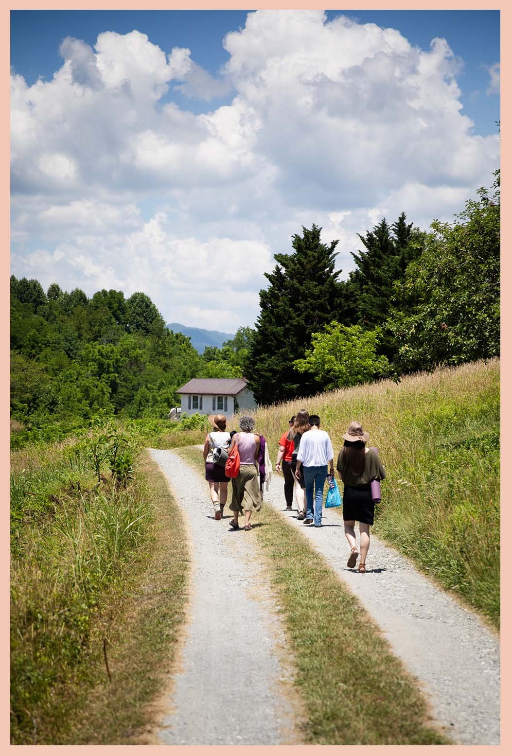 A group of people walking on a gravel country road surrounded by grass and trees with a house in the background and a partly cloudy sky above.