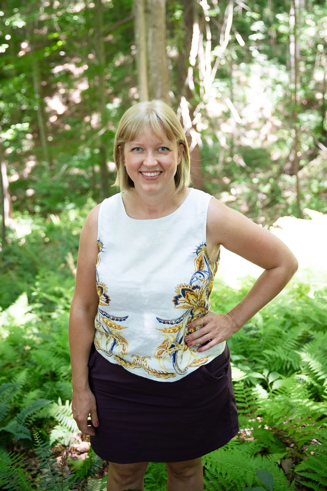 A woman standing in a lush green forest with ferns, smiling at the camera, wearing a sleeveless white top with colorful patterns and a dark skirt.