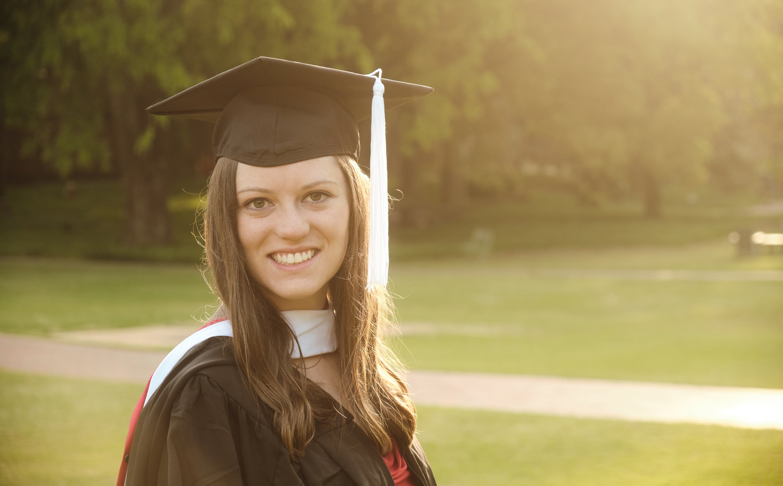 A young woman smiling in a graduation cap and gown, outdoors in a park, with sunlight filtering through trees in the background.