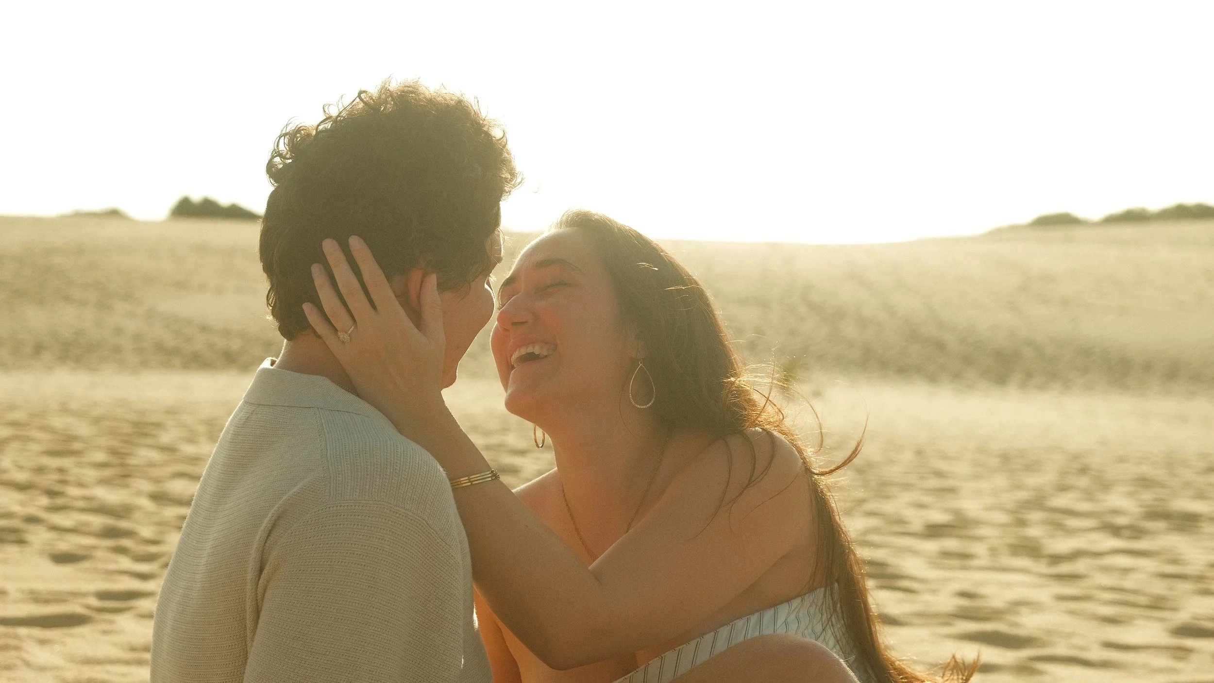 A couple smiling and embracing on a sandy beach at sunset.