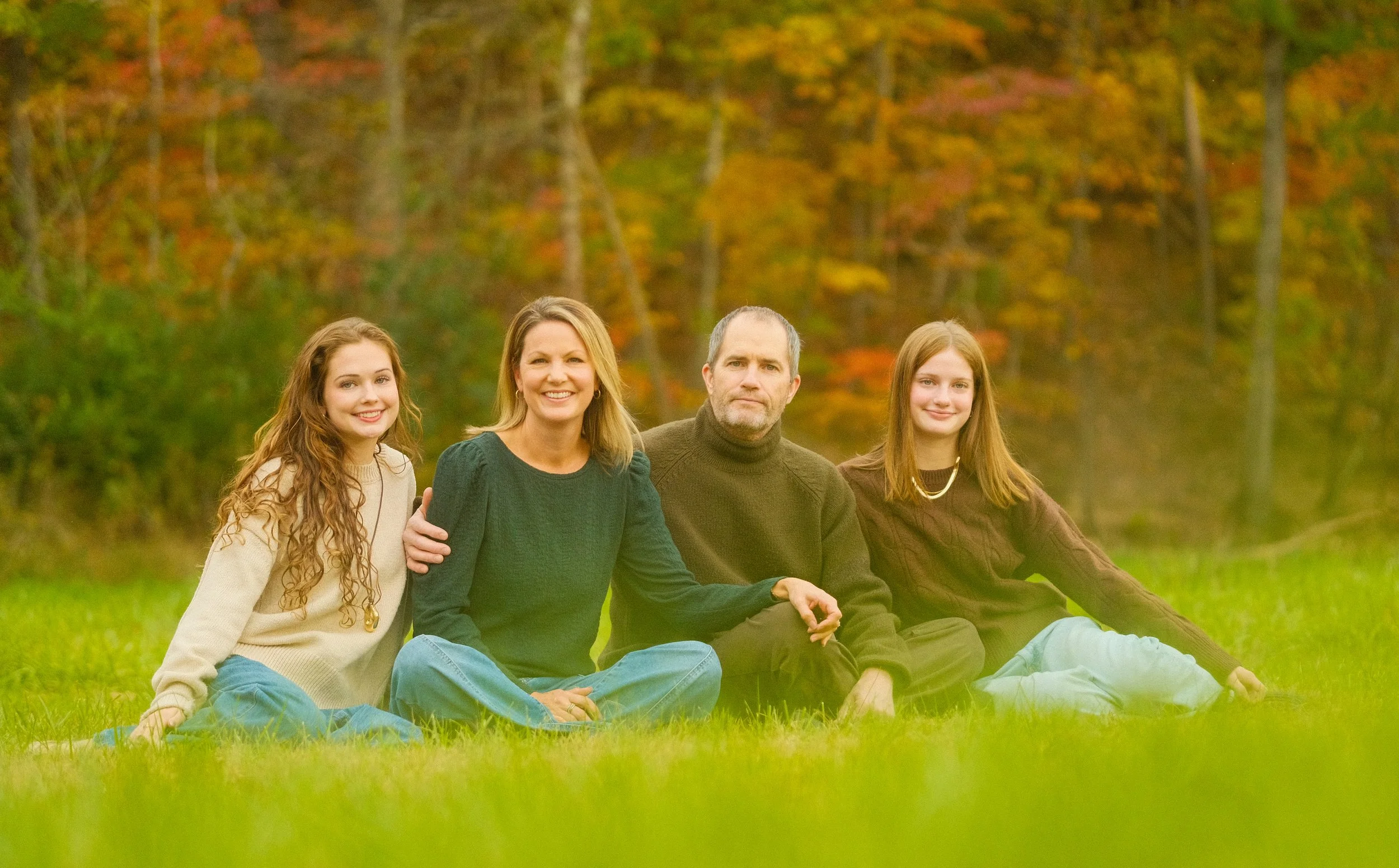 A family of four sitting on grass outdoors in a park with autumn foliage in the background.