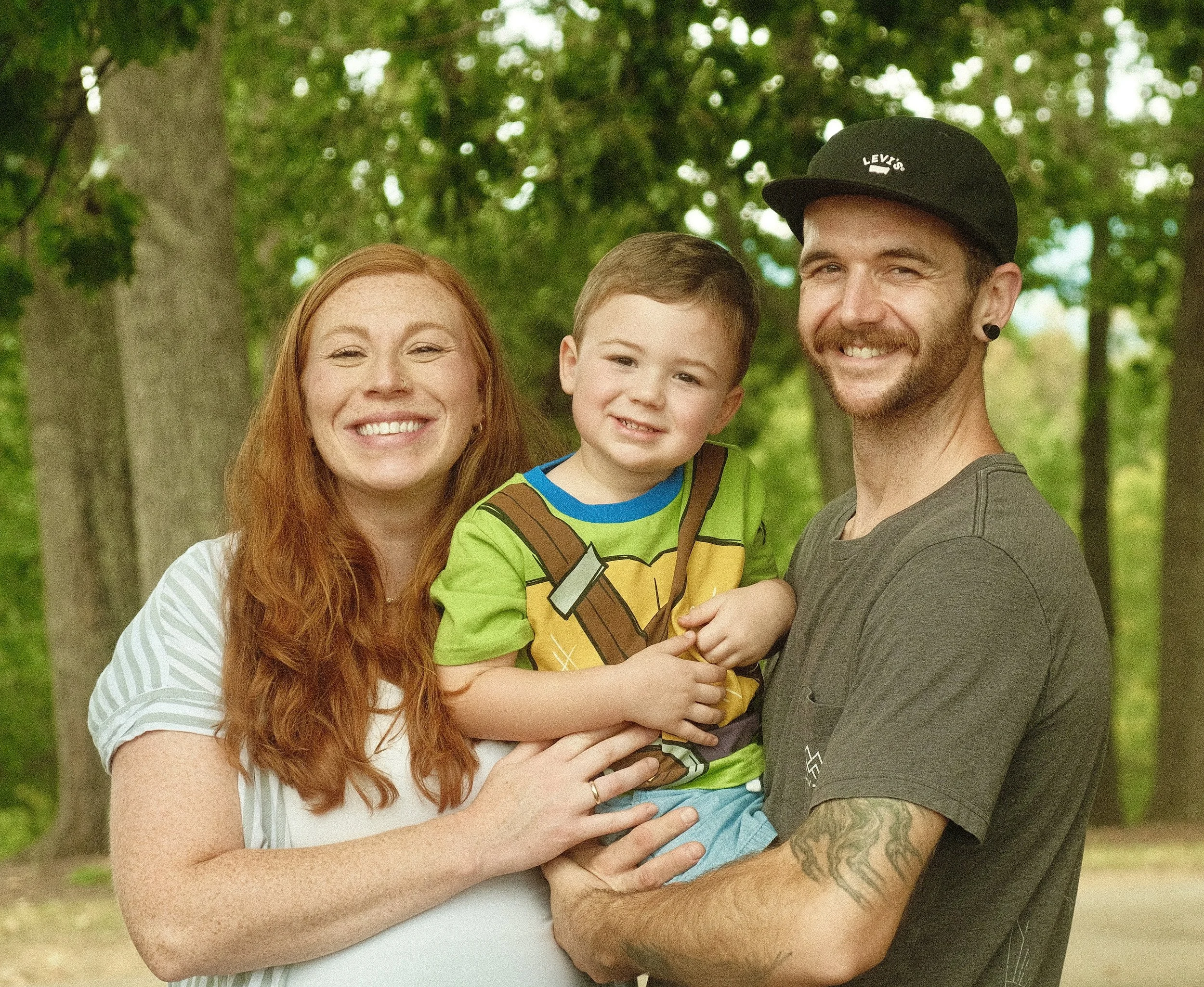 Family portrait of a smiling mom and dad holding their child outdoors in Salem, Virginia, surrounded by trees and natural greenery.