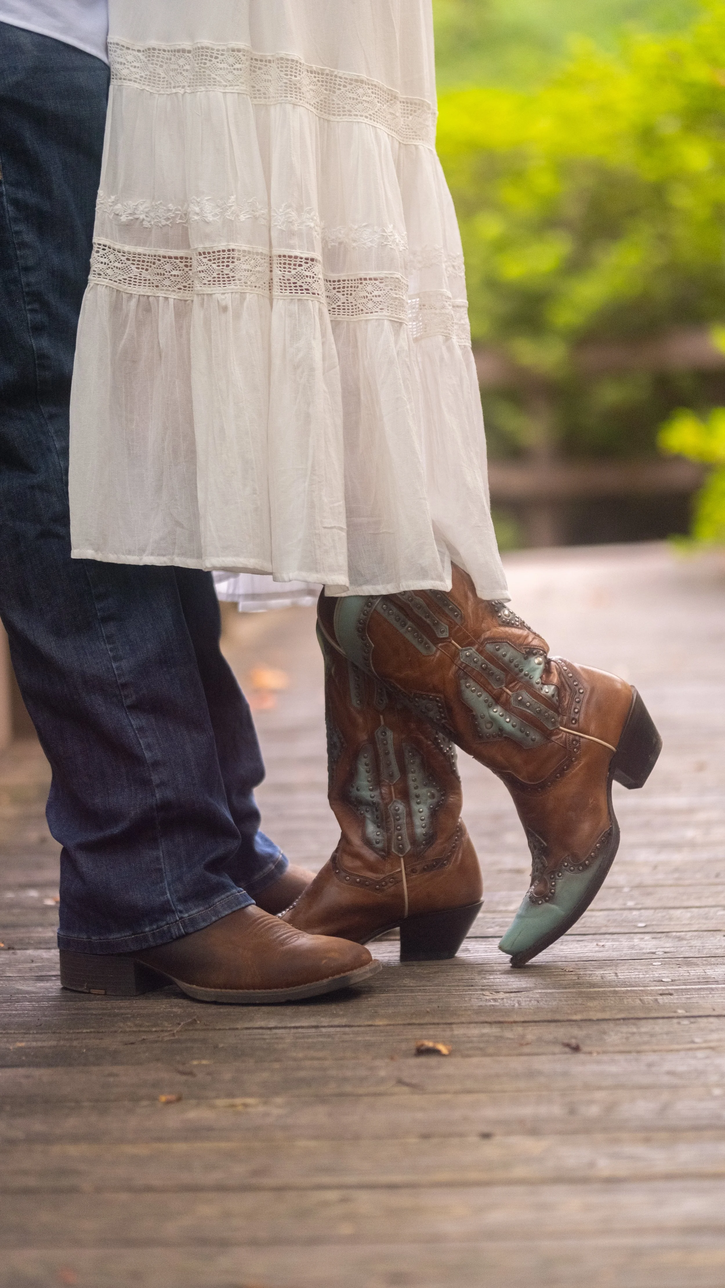 A photo of a couple's cowboy boots with a bridge and foliage in the background