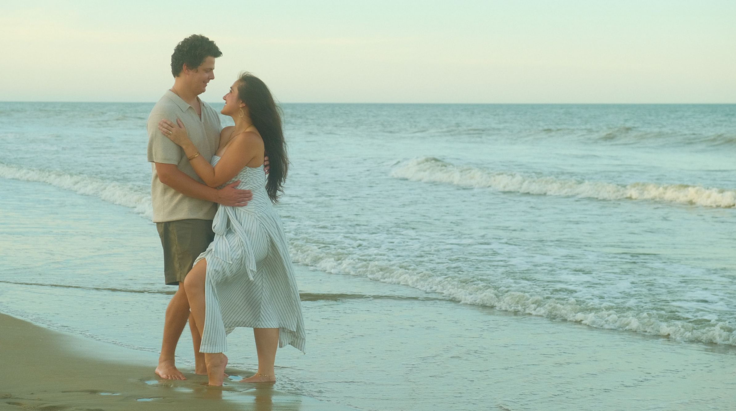 A couple smiling and embracing on a sandy beach at sunset with waves in the background.