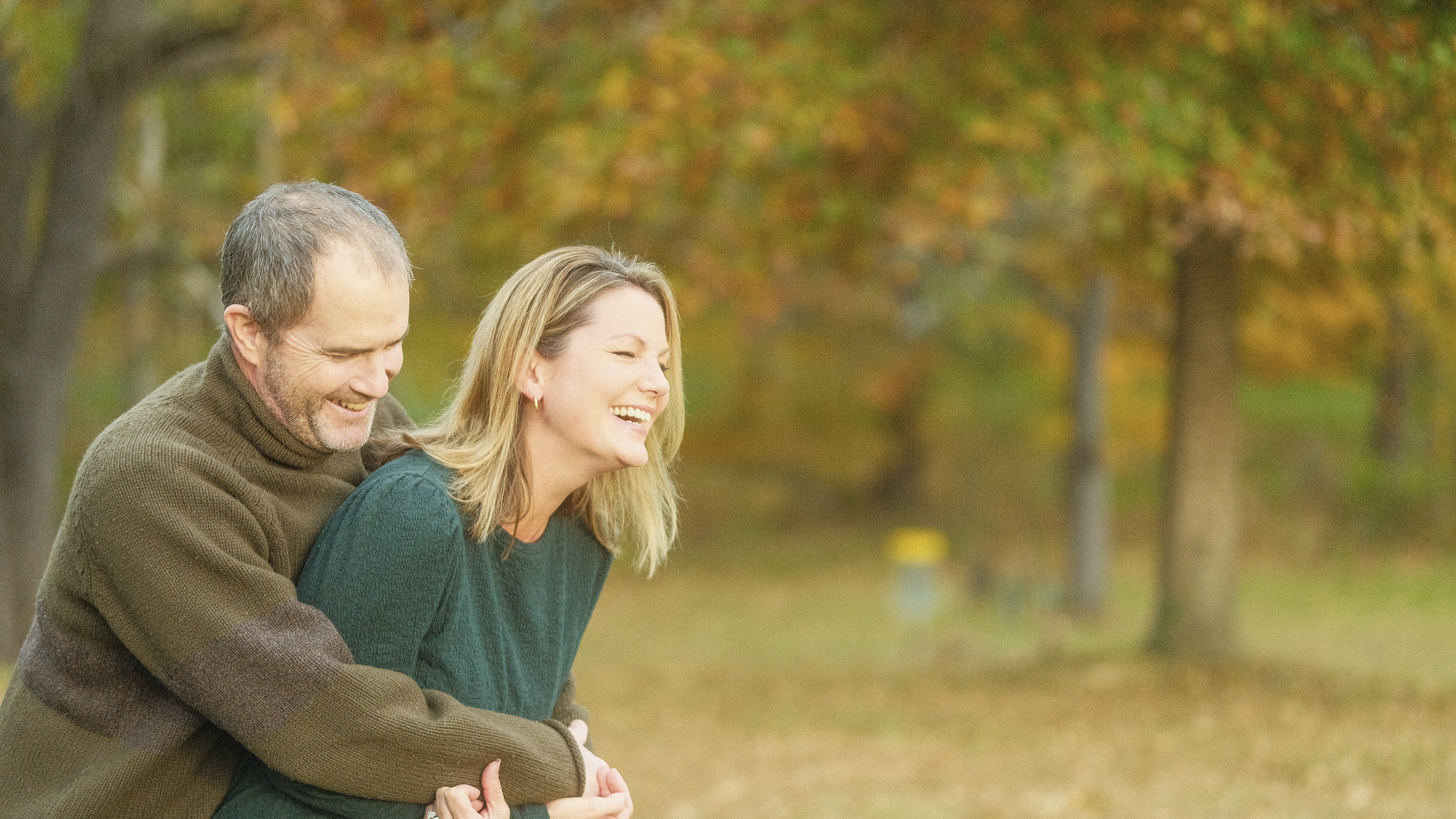 A man and woman outdoors in autumn, smiling and enjoying a moment together, with trees and fall foliage in the background.
