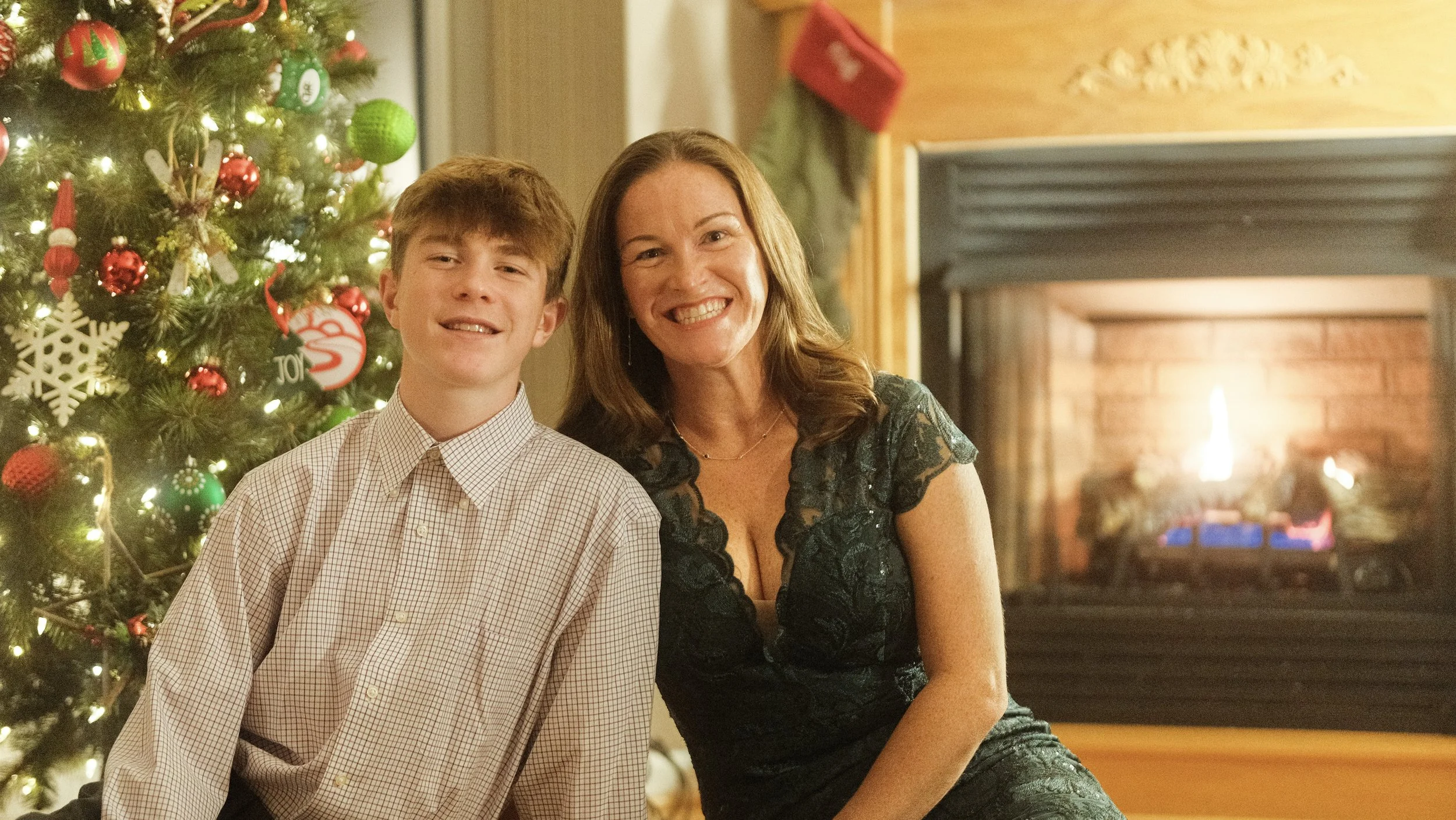 A smiling woman and teenage boy sitting together in front of a decorated Christmas tree with ornaments, lights, and a fireplace in the background.