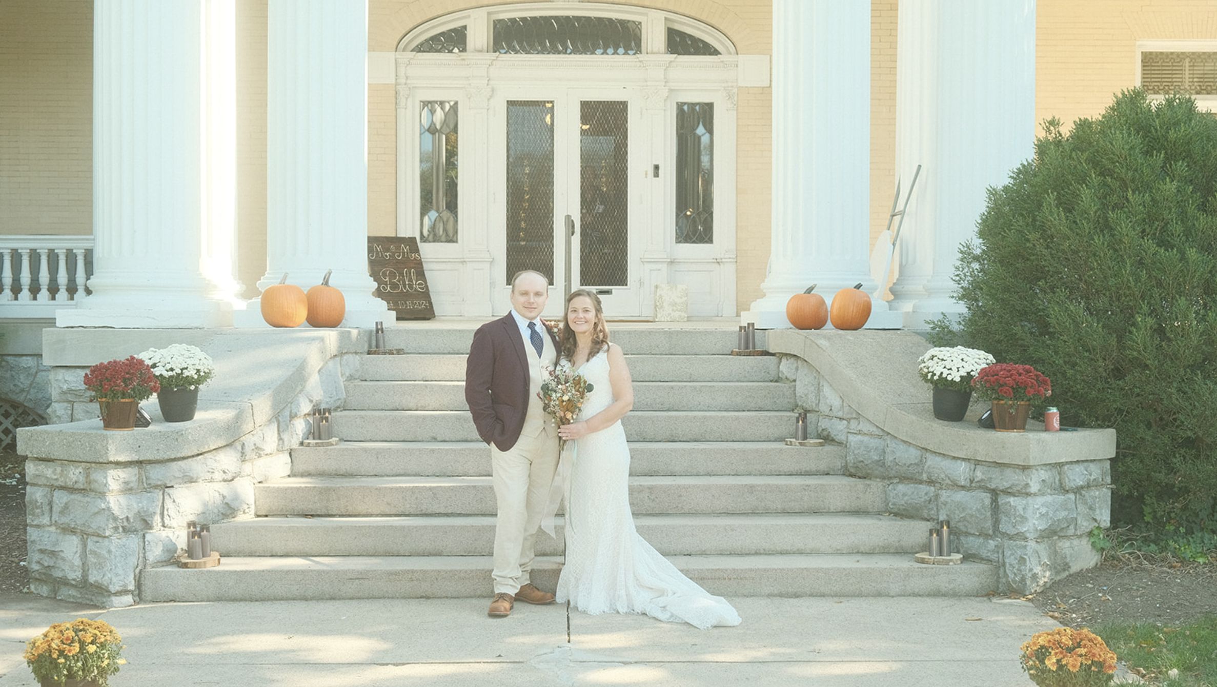 Wedding portrait of couple in front of a mansion's staircase. Woman is wearing a long white bridal gown and holding dried flowers. The male is in a suit and tie.