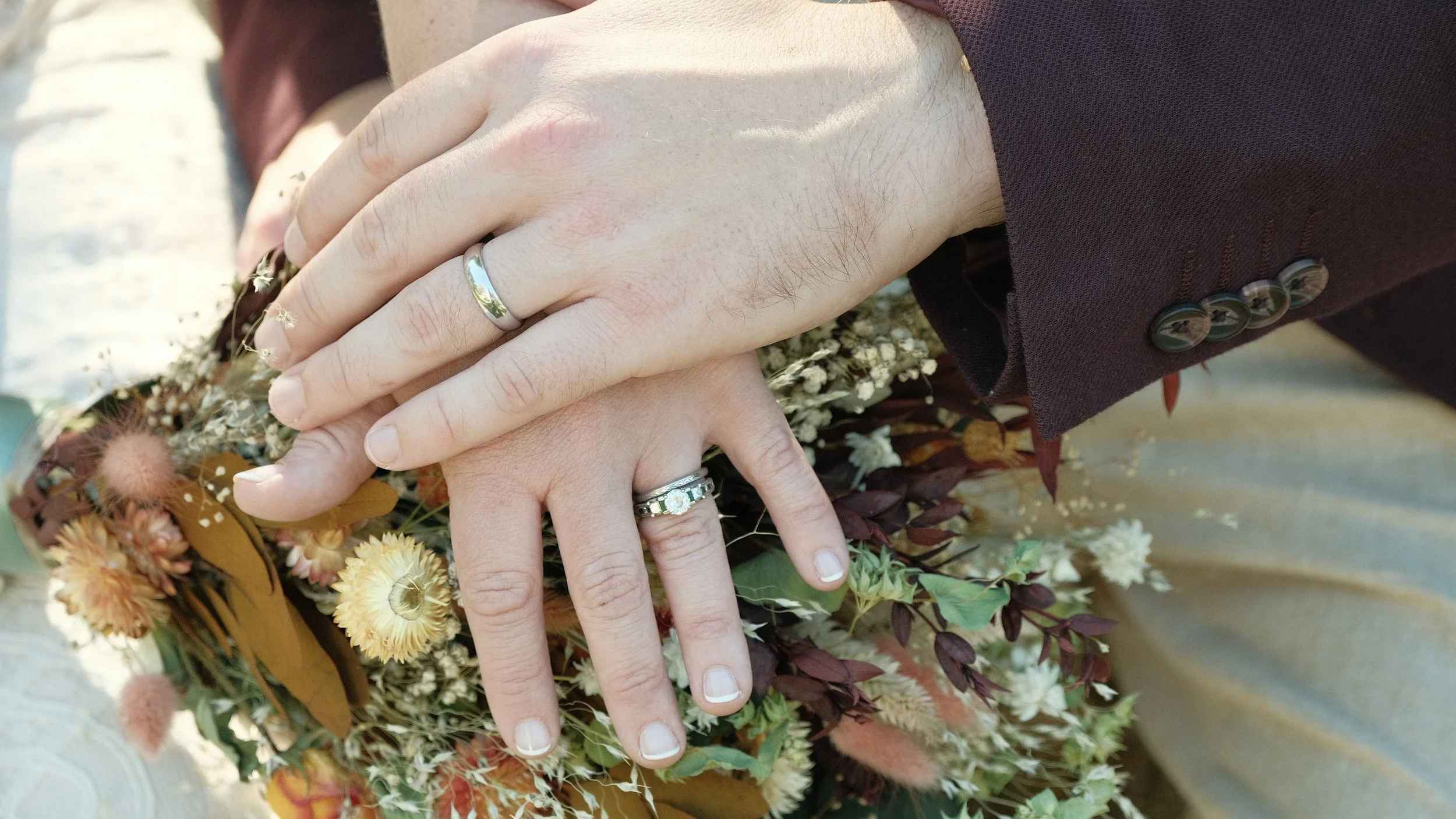 Close-up of two hands with wedding rings resting on top of a bouquet of flowers, including yellow, white, and orange blossoms, on a light-colored surface. One person is wearing a dark suit jacket.