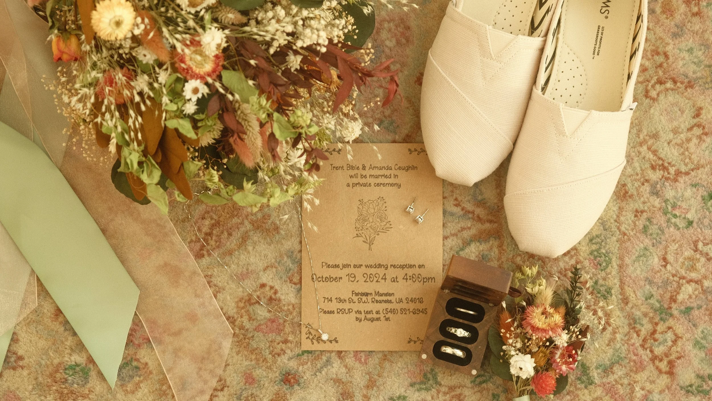 A wedding announcement card with dried flower bouquets, white slip-on shoes, wedding rings, and small earrings on a floral patterned carpet.
