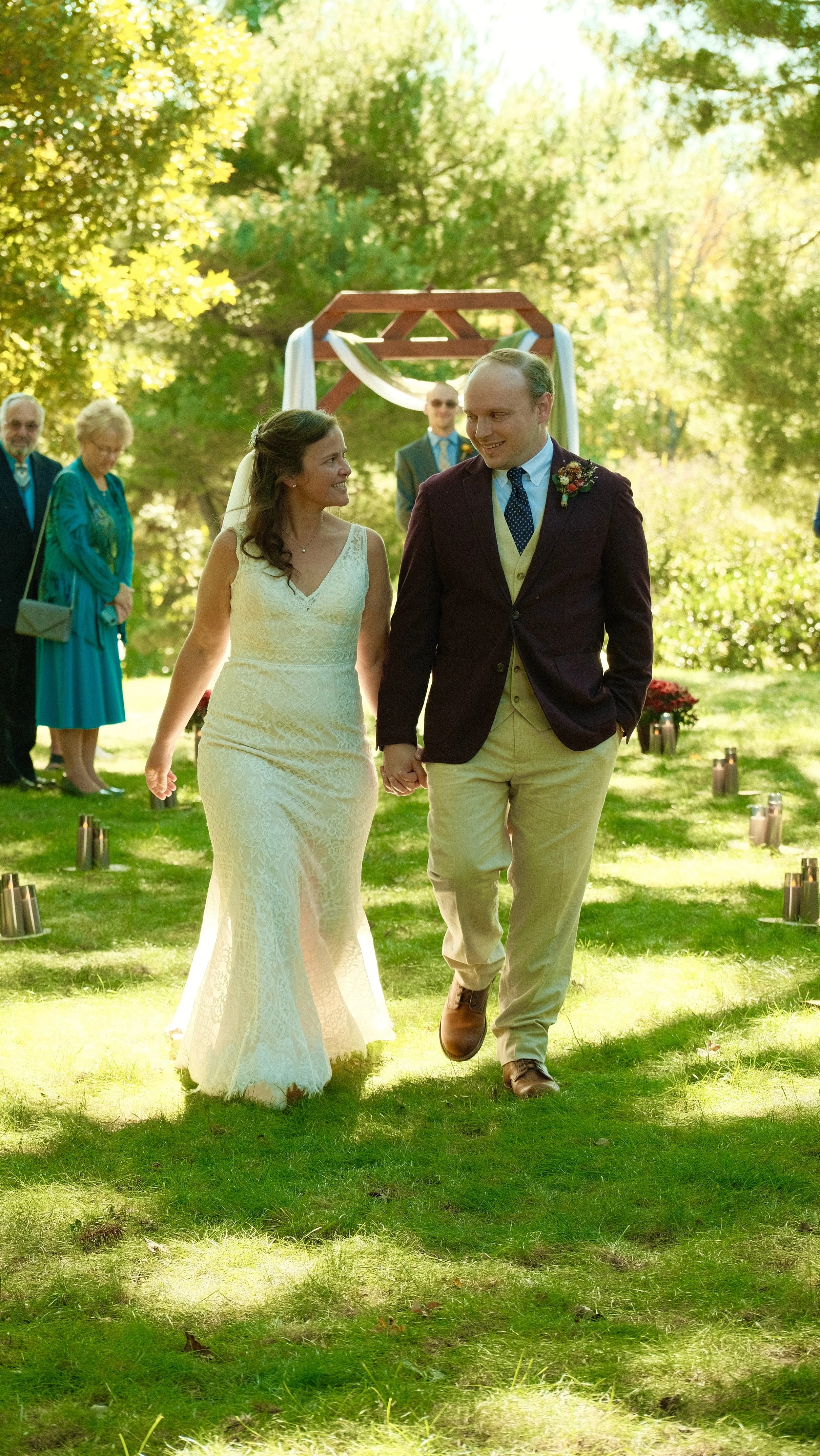 A bride and groom walking hand in hand outdoors during a wedding ceremony, with family and friends in the background under trees on a sunny day.