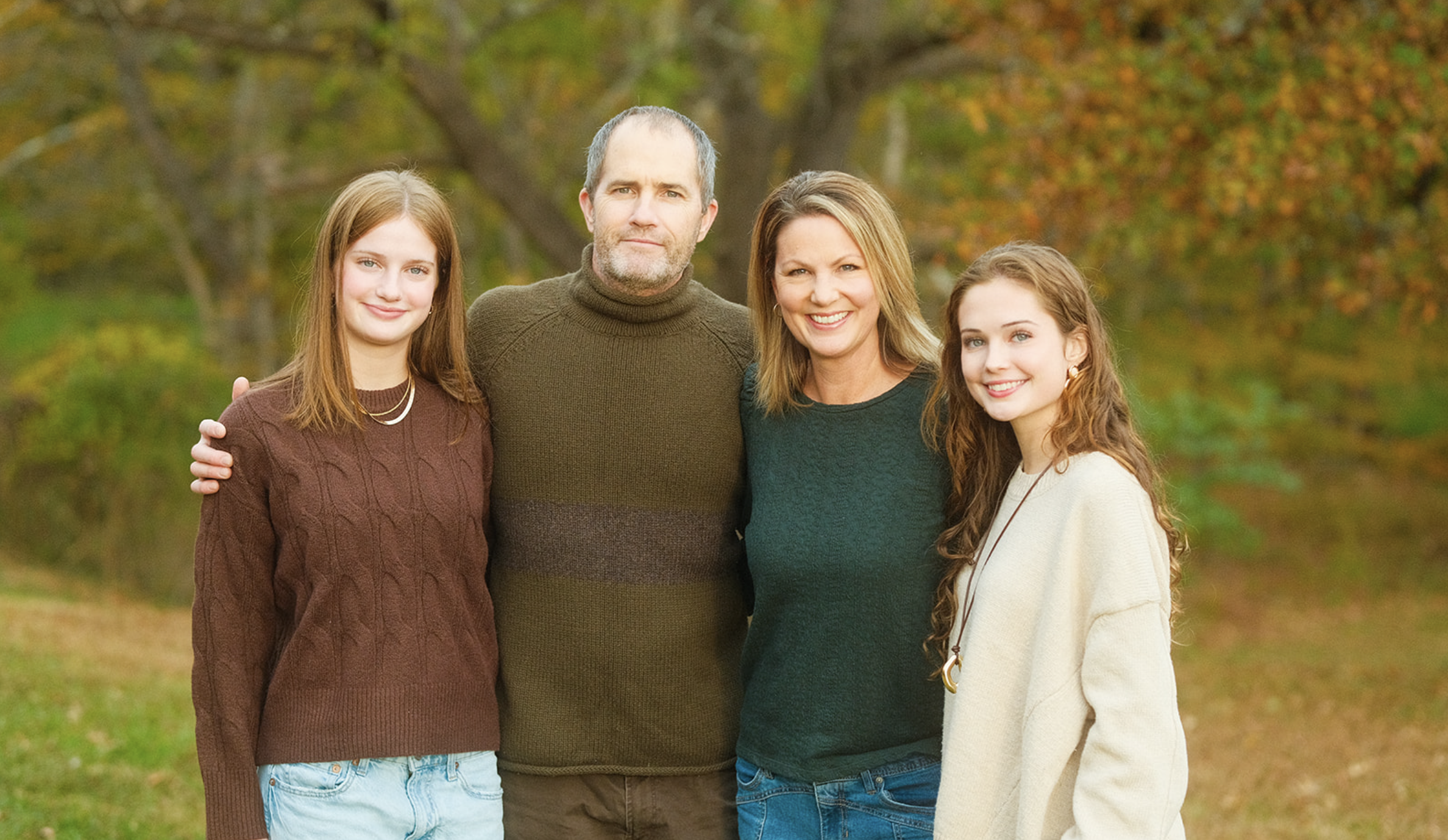 Family portrait of a mom, dad, and two daughters smiling with fall leaves in the background.