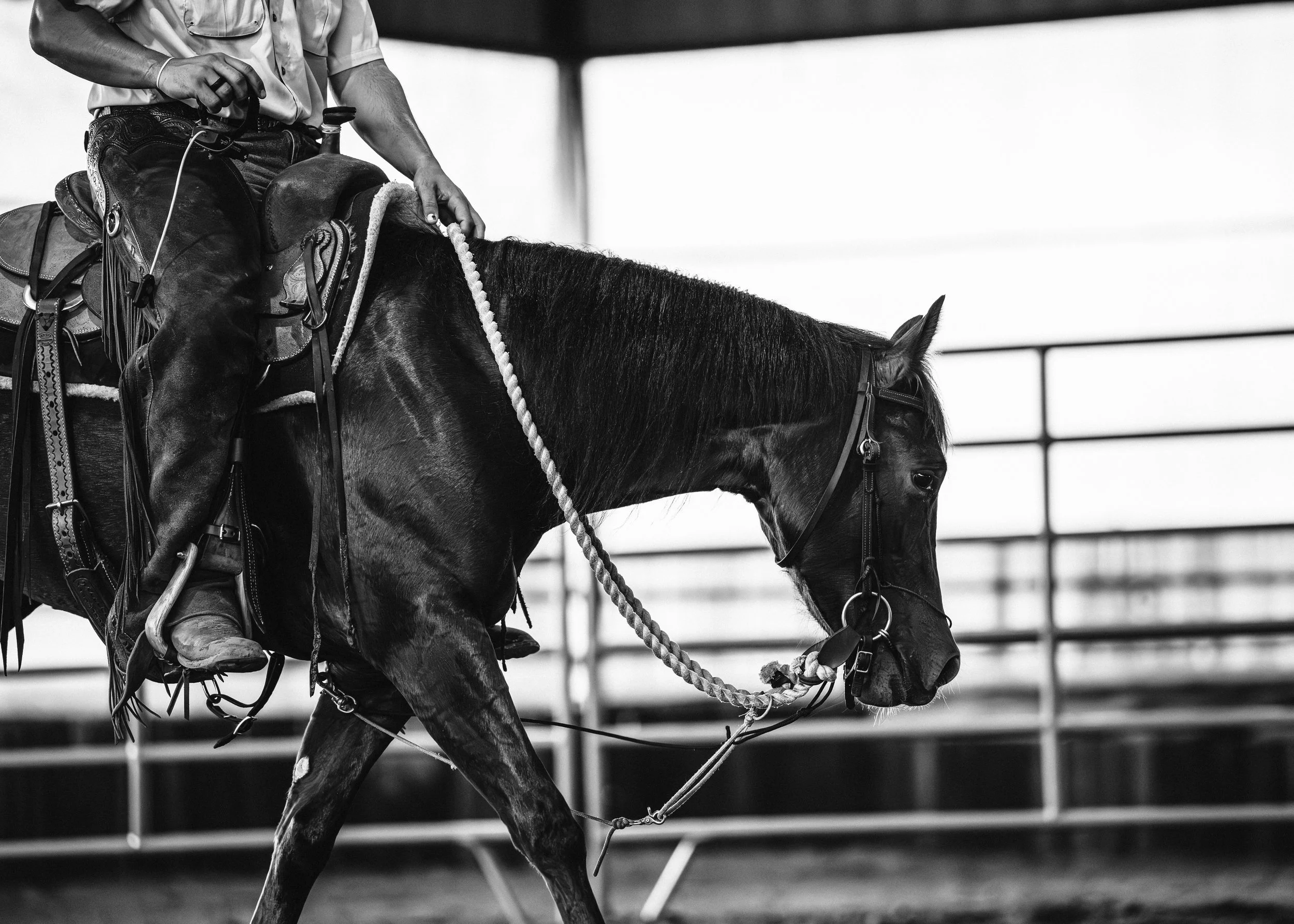 A black-and-white photograph of a person riding a horse indoors, with a railing visible in the background.