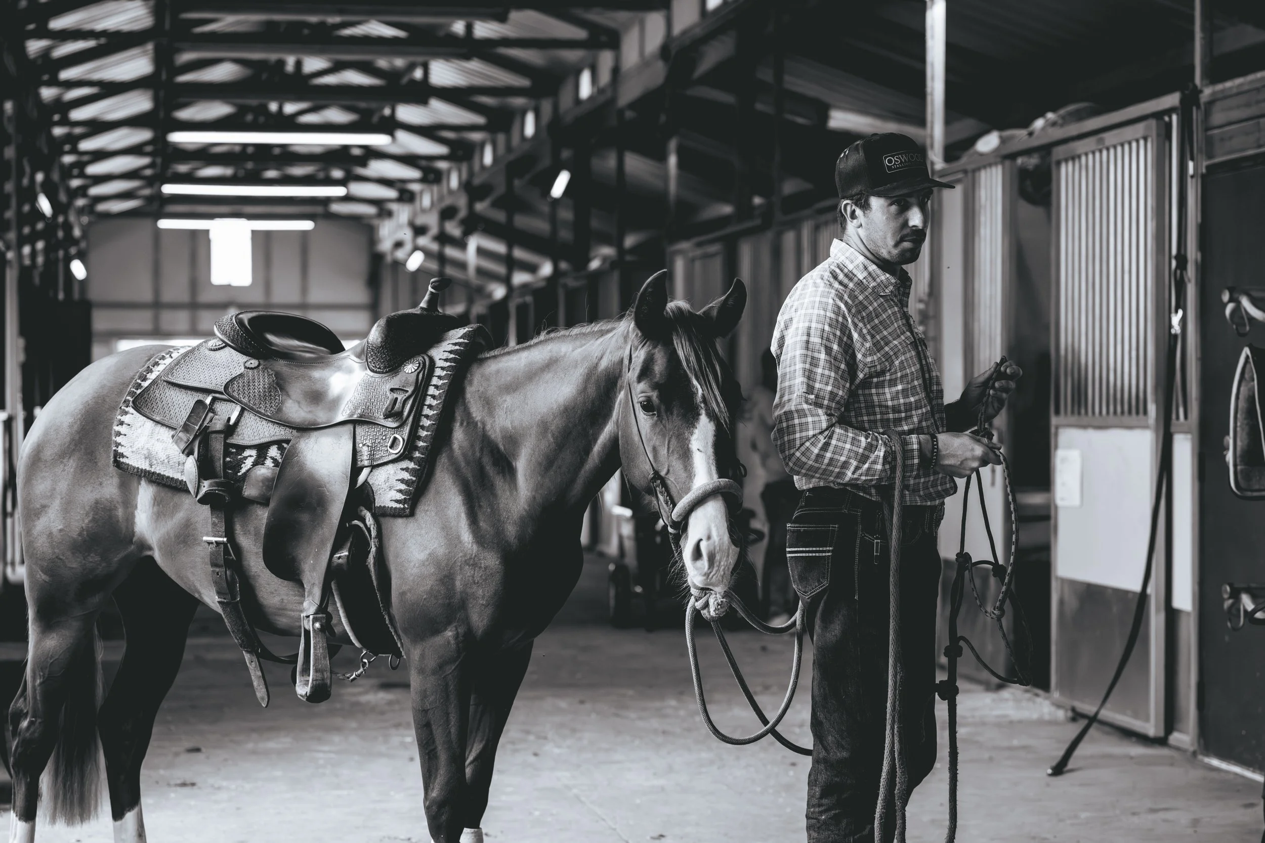 A man holding the lead of a saddled horse inside a rustic barn.