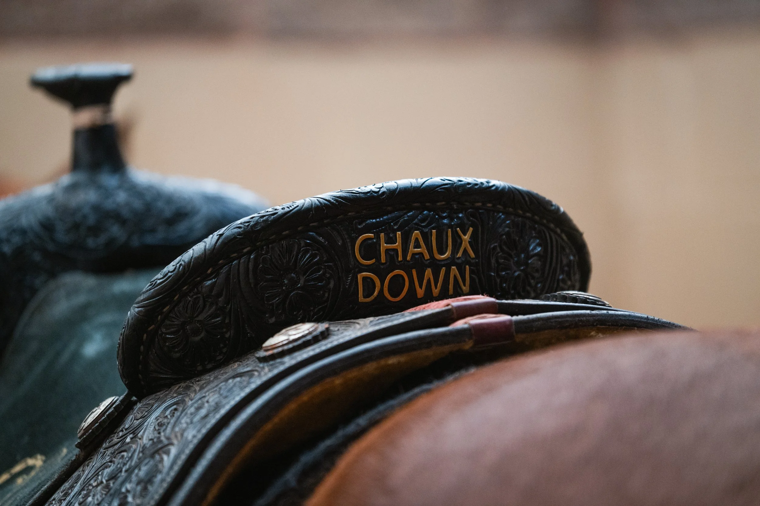 Close-up of a cowboy saddle with decorative tooling, with a leather strap displaying the words 'Chaux Down' in yellow lettering.