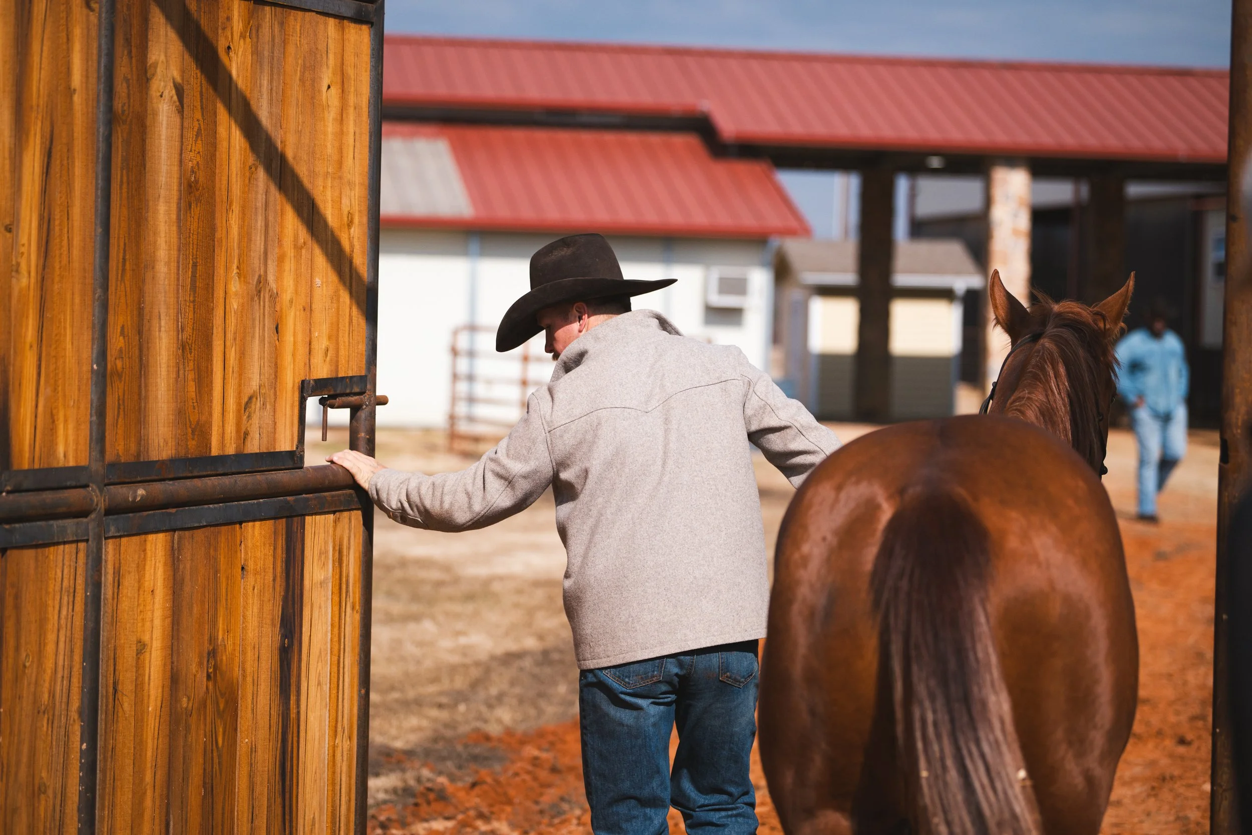 A man in a cowboy hat and gray jacket leads a brown horse into a stable at a ranch with wooden structures and other people in the background.