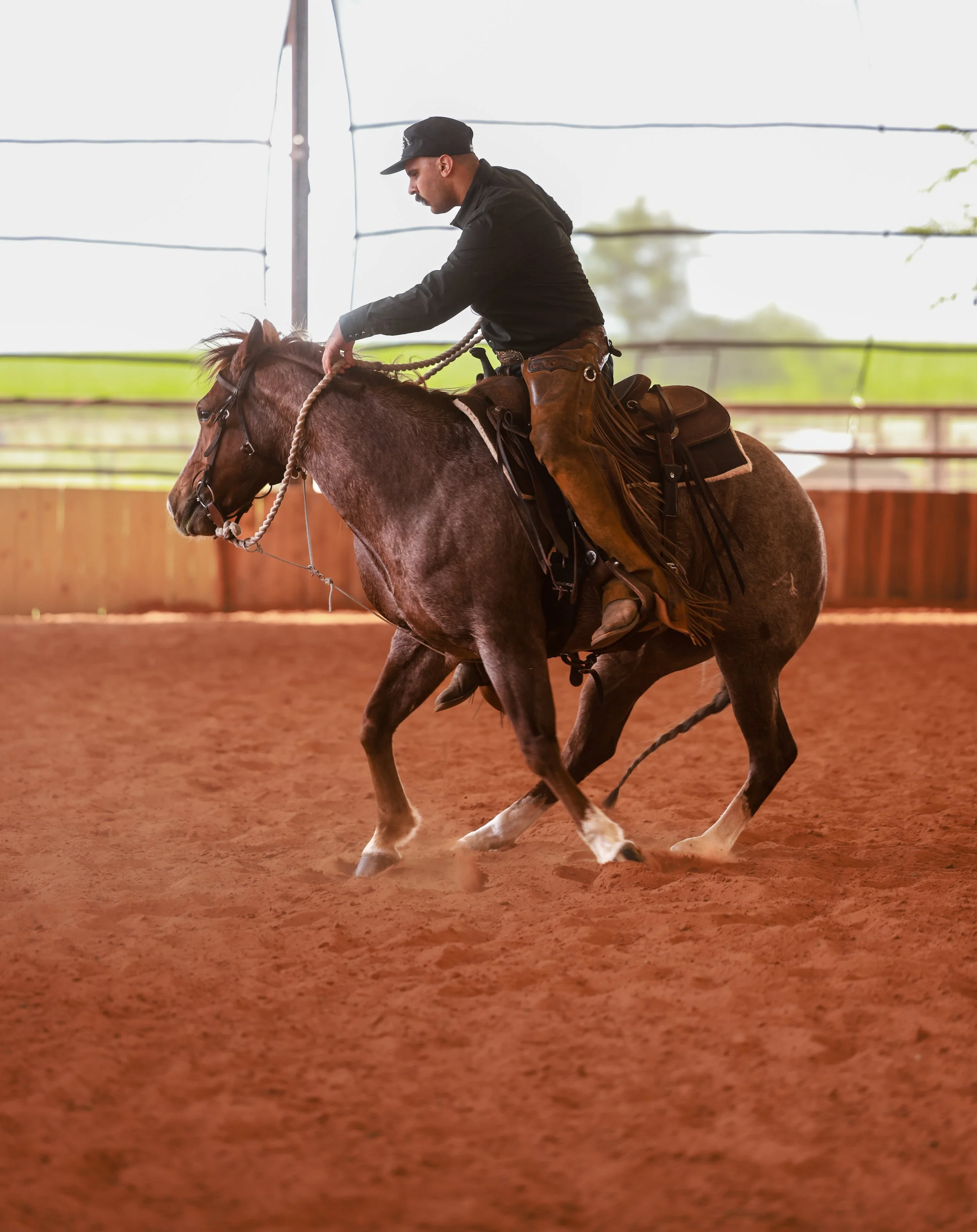 Person in cowboy hat and gray jacket riding a brown horse in an outdoor arena with a wooden fence.