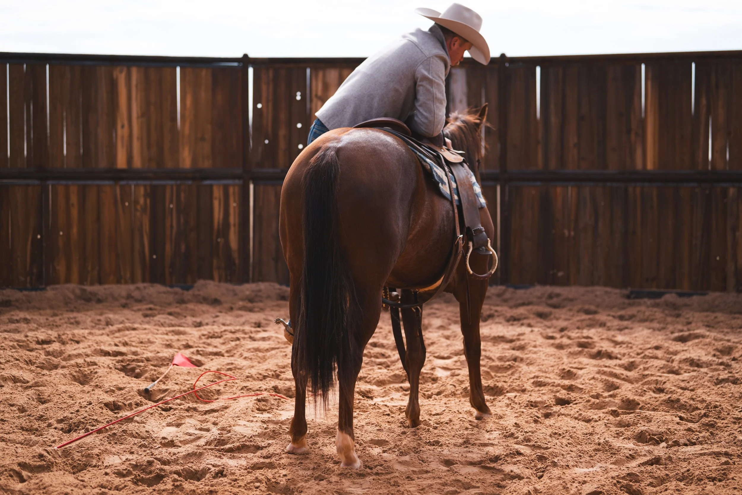 Person in cowboy hat and gray jacket riding a brown horse in an outdoor arena with a wooden fence.