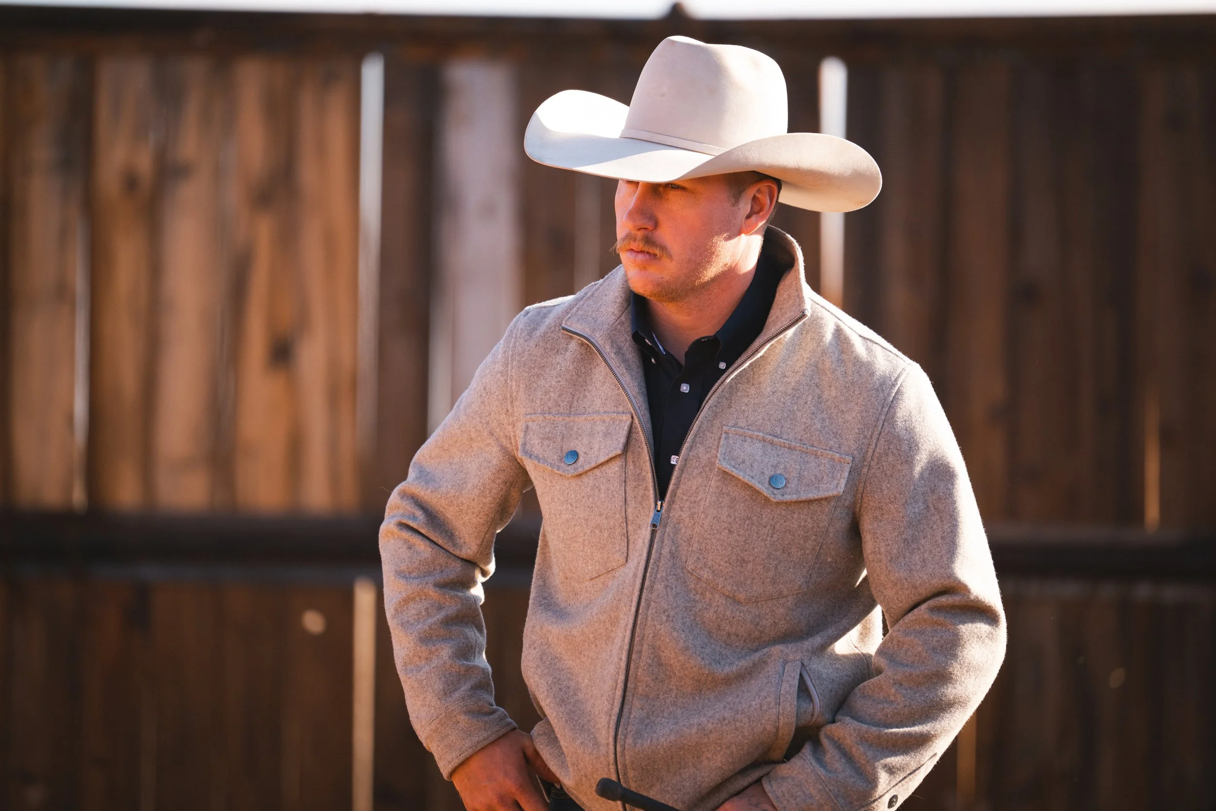 A man wearing a light-colored cowboy hat, a gray zip-up jacket with pockets, and standing outdoors near a wooden fence.