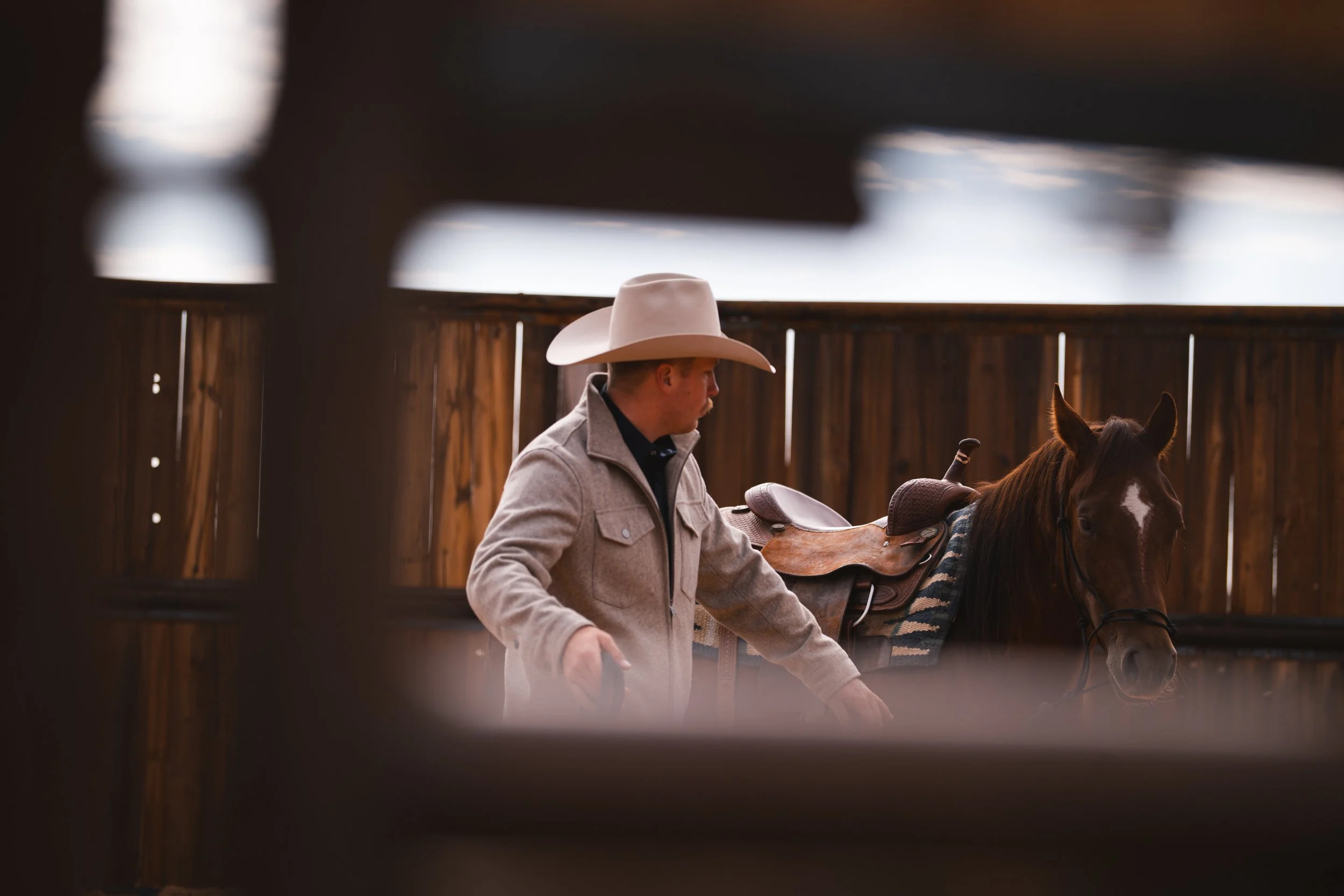 A man wearing a cowboy hat and a beige jacket standing next to a brown horse with a saddle, inside a wooden fenced area.