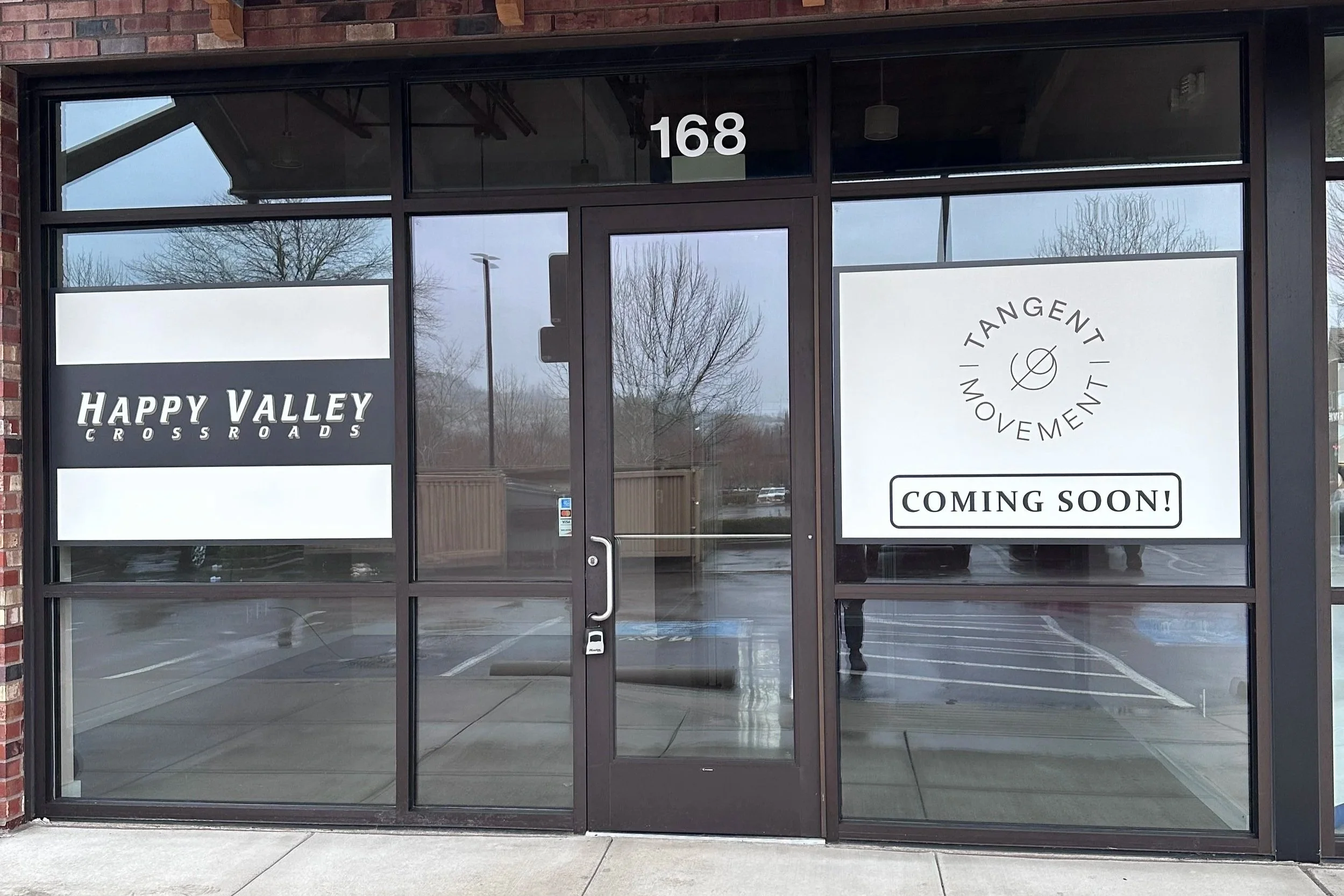 Storefront window with signage announcing a new business TANGENT MOVEMENT coming soon, and another sign for HAPPY VALLEY CROSSROADS.