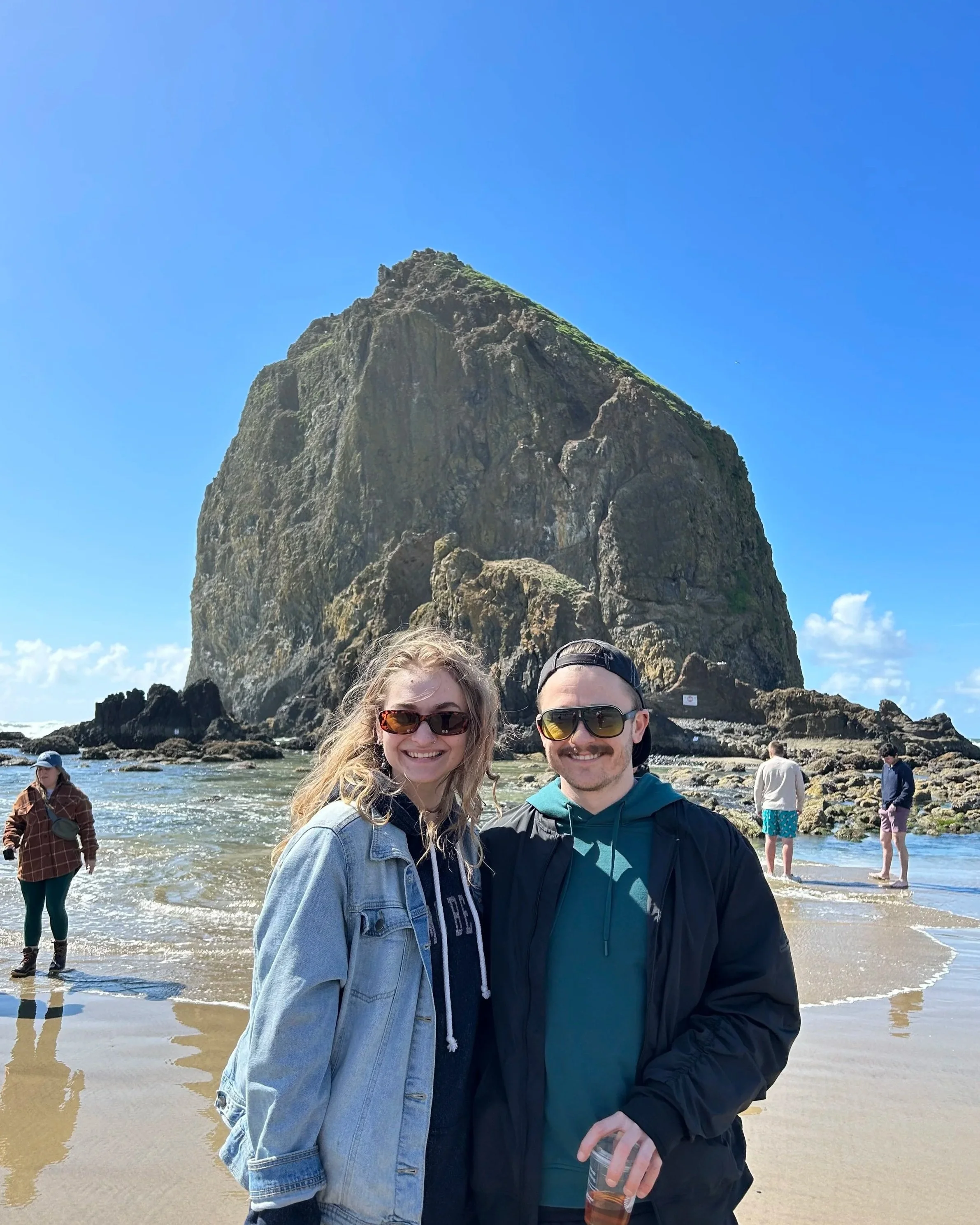 Two smiling people standing on a beach with a large sea stack in the background. The woman has wavy blonde hair, sunglasses, and a denim jacket. The man has a beard, sunglasses, a backwards cap, and a dark jacket. There are other people exploring in the background under a clear blue sky.