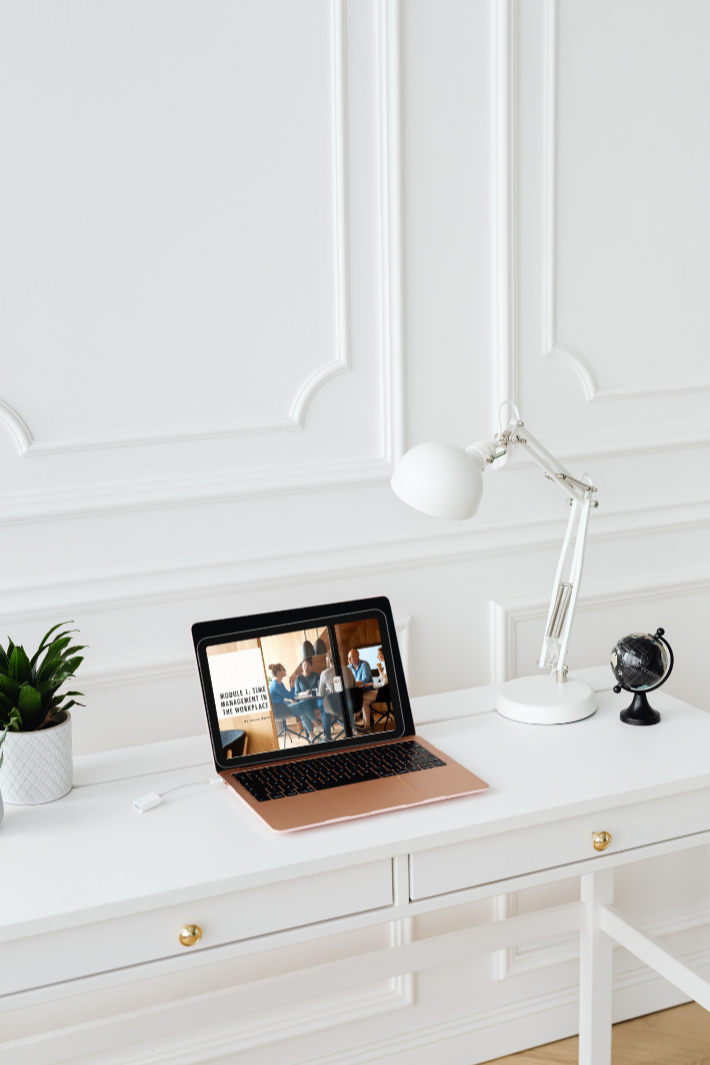White desk with a rose gold laptop displaying a meeting, a white study lamp, a small globe, and a potted plant against white paneled wall.