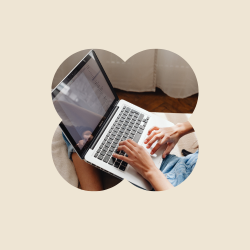 A person using a laptop on their lap, sitting on a couch with beige background and floral curtains.