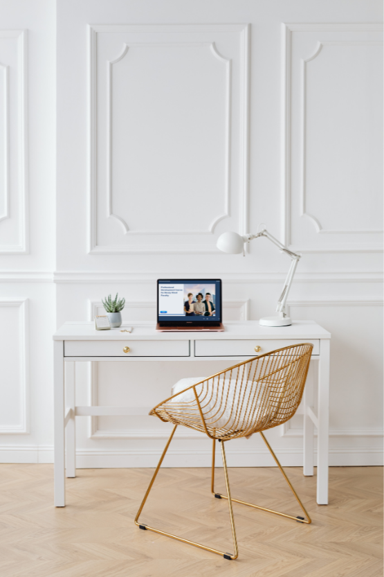 A white desk with two drawers and golden knobs, a laptop displaying a presentation, a small potted plant, and a white adjustable desk lamp. A gold wire chair with a white cushion is in front of the desk. The backdrop features white wall paneling with decorative molding.