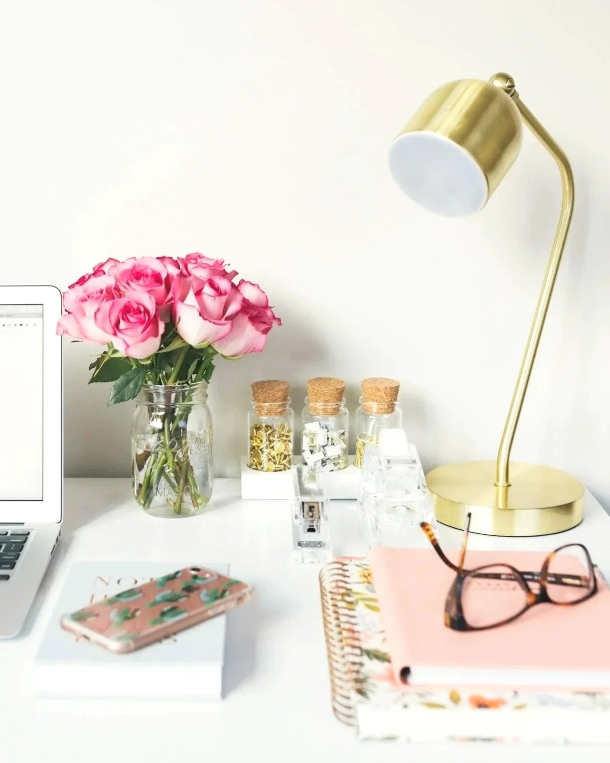 A desk with a laptop, a glass jar of pink roses, three small glass jars with cork lids containing gold and white decor, a gold desk lamp, a pink patterned notebook with glasses on top, and a planner.