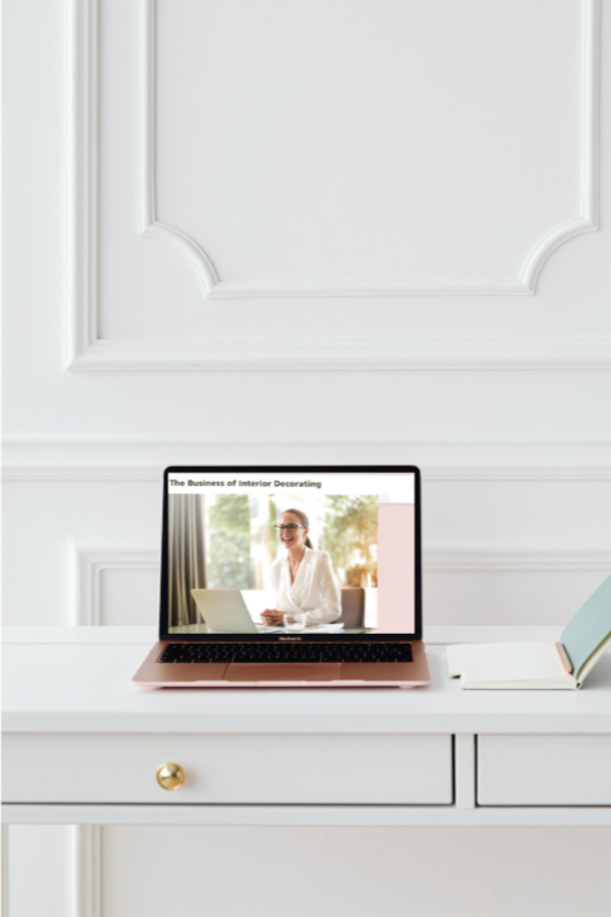Laptop on white desk displaying a woman in glasses and a white blazer, with a window and trees in the background, next to a closed book.