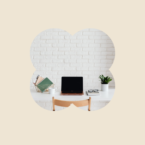 Desk with open laptop, reading glasses, white mug, potted plant, and books against a white brick wall