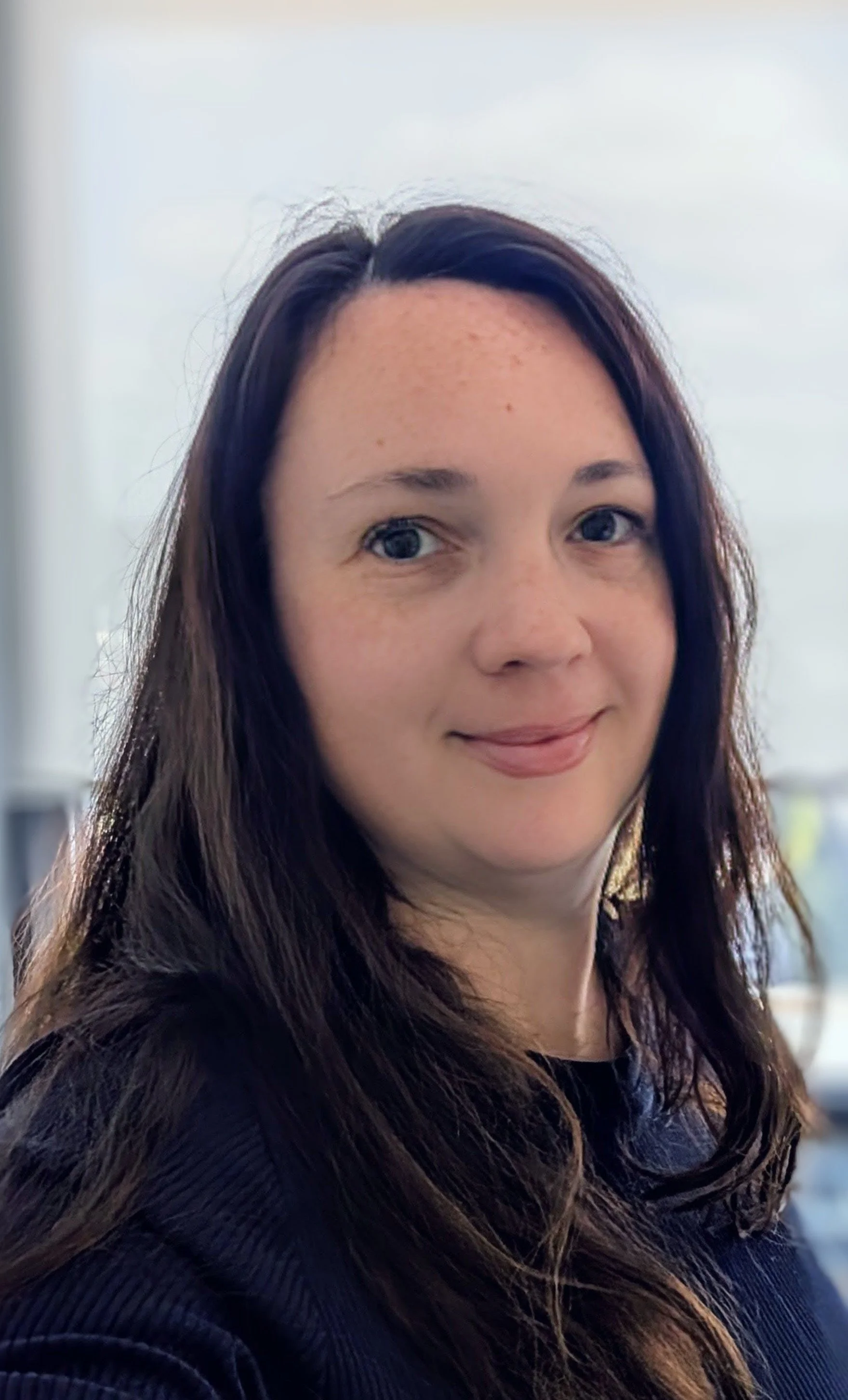Portrait of a woman with long brown hair, smiling softly, indoors with a blurred background.