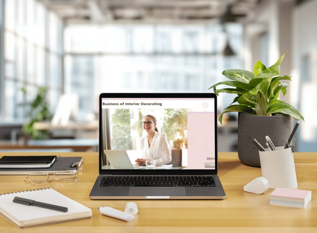 Laptop on a wooden desk displaying a woman sitting at a desk, with a title about interior decorating business. Desk has a notebook, pen, glasses, sticky notes, a plant in a pot, a cup with pens, a white bulb, and a small white device. Windowed office background.